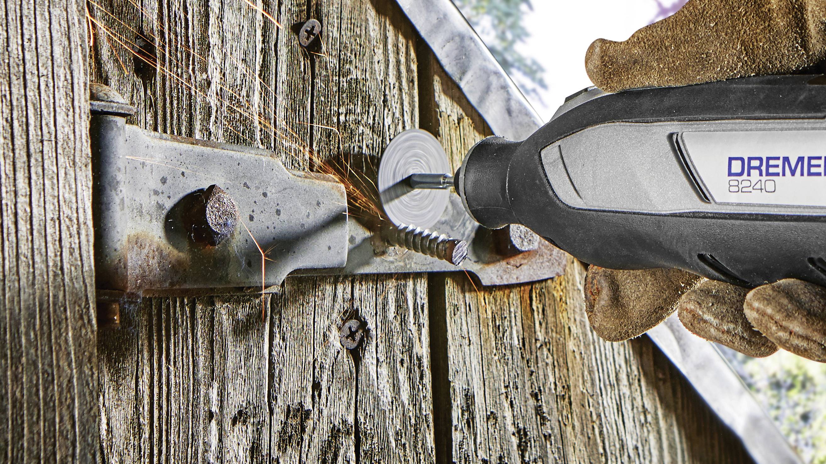 A person is cutting a rusty metal hinge on a wooden gate using a Dremel tool. Sparks are flying as they cut.