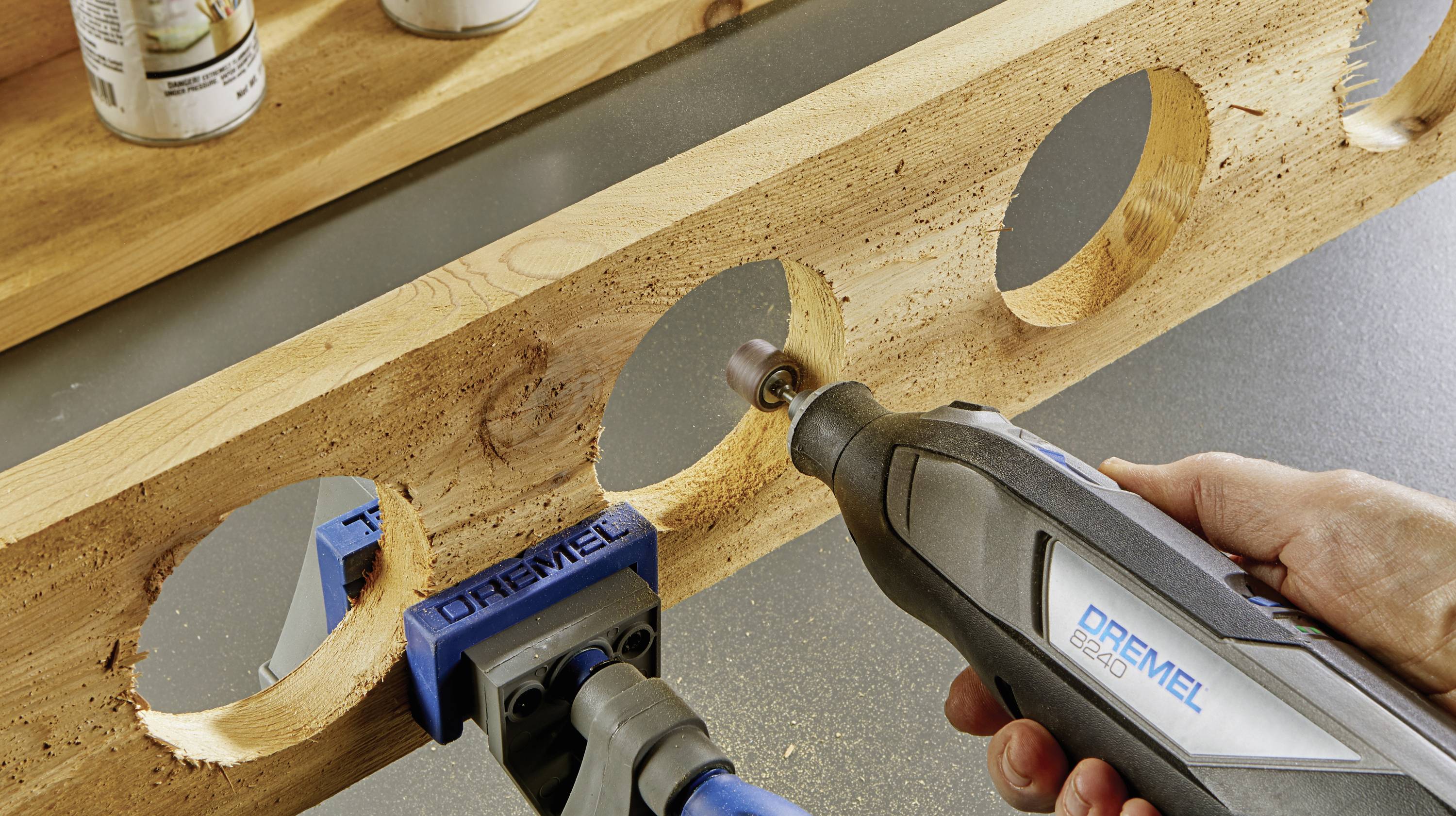 A person is sanding the edges of large circular holes in a wooden board, which is secured in a vice, using a Dremel tool.