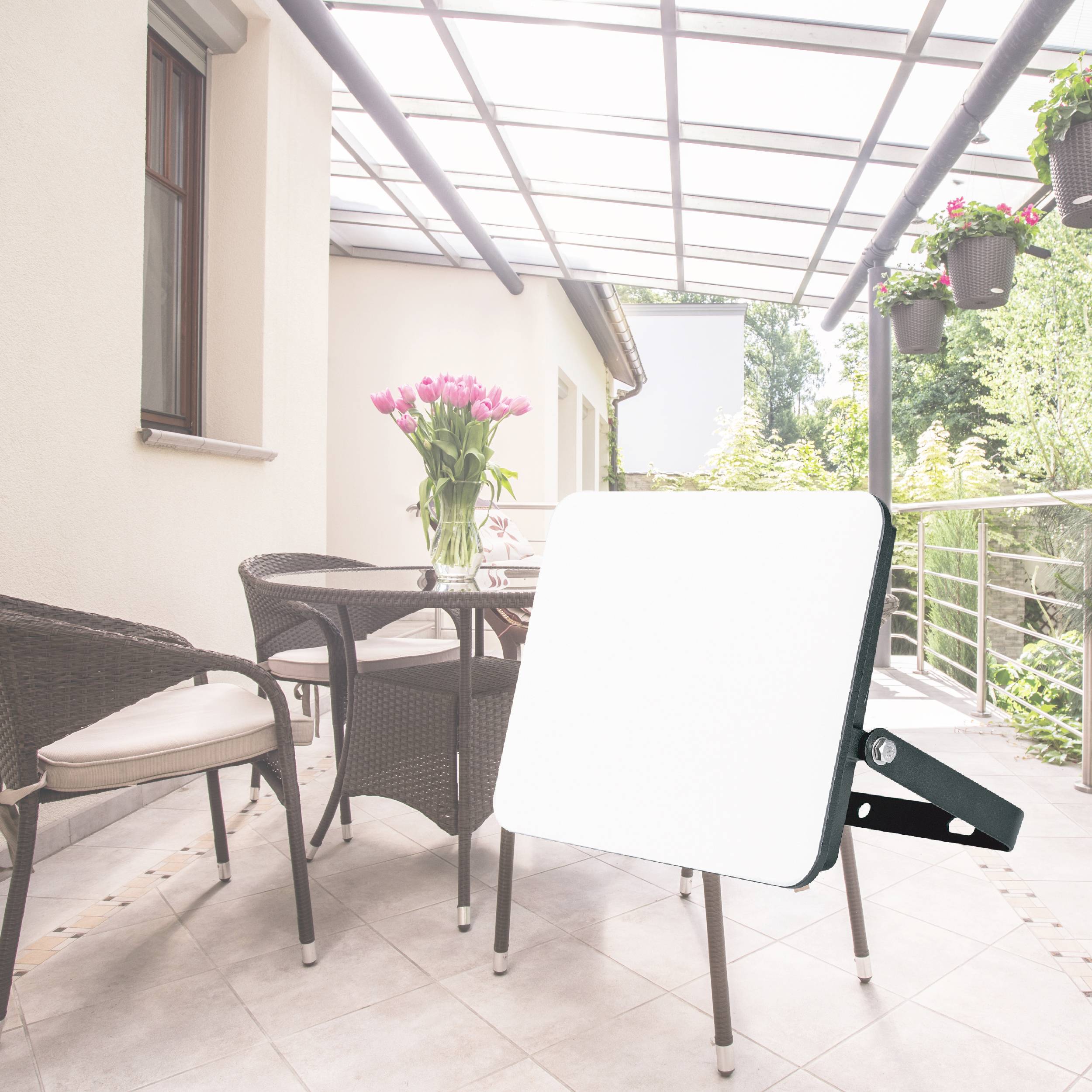 A modern terrace with a glass roof, two chairs and a round table. Flowers are visible in the background.