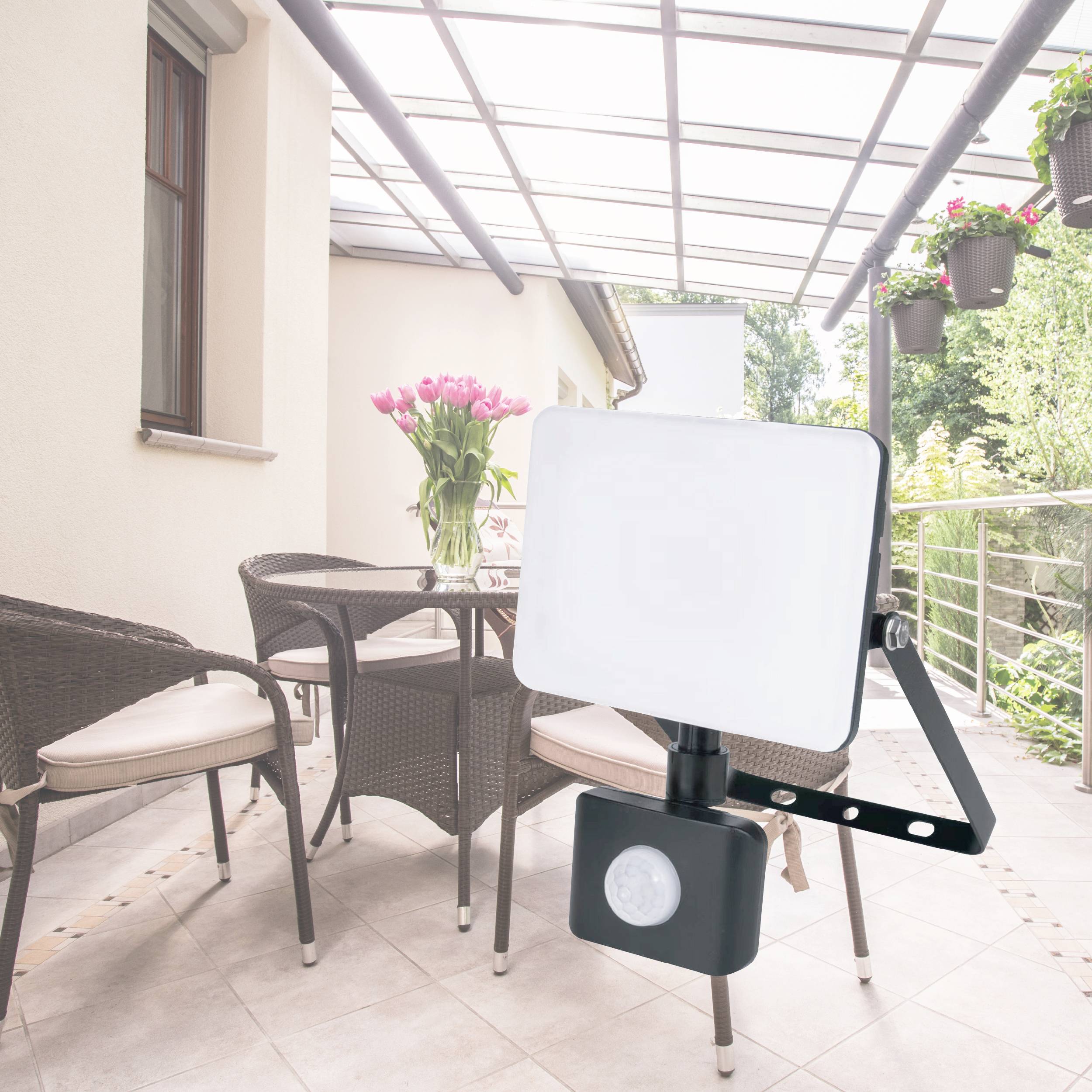 A bright outdoor spotlight with a motion sensor is installed on a covered terrace. Furniture and plants are visible in the background.