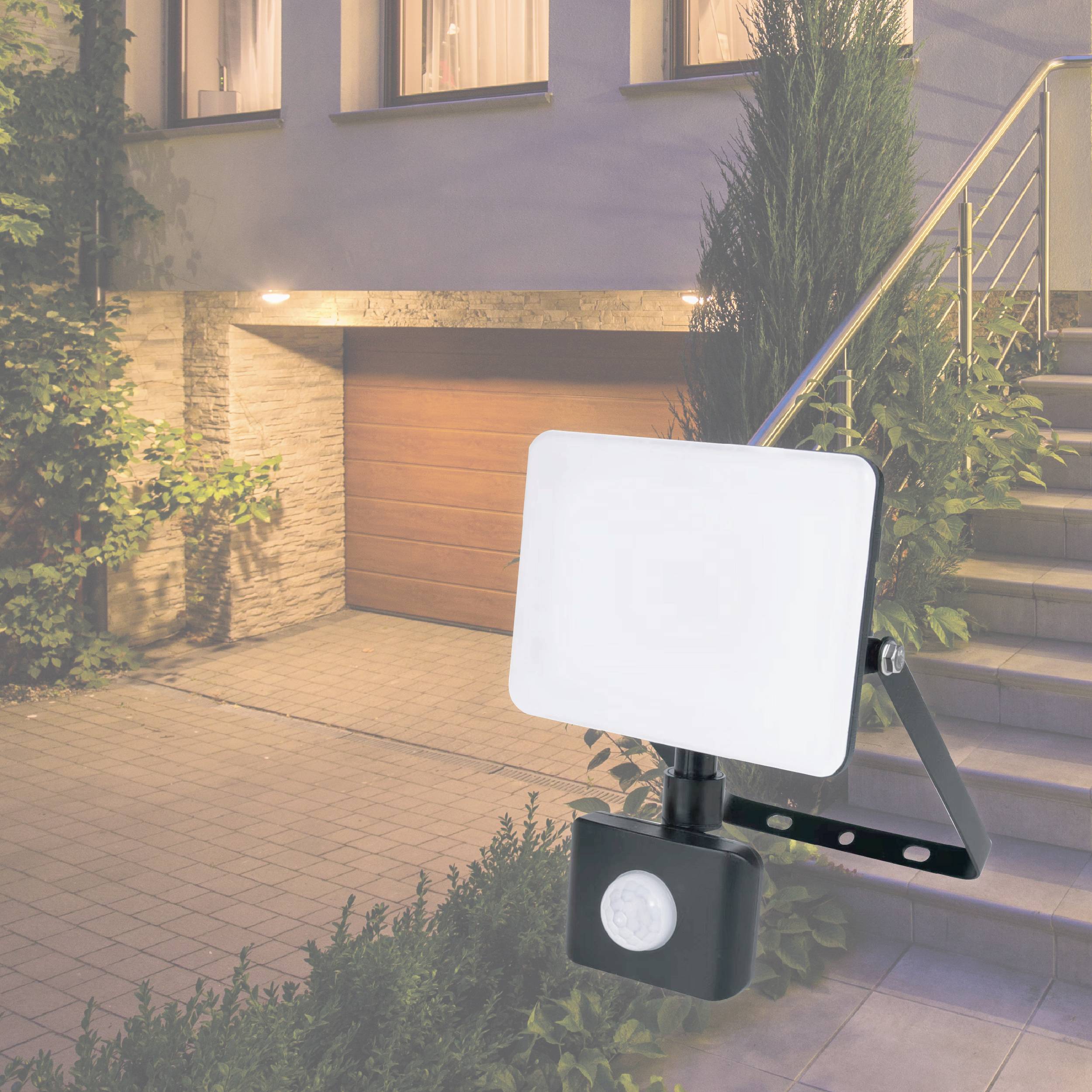A motion sensor with an attached LED floodlight is installed in front of a garage and illuminates the paved courtyard.