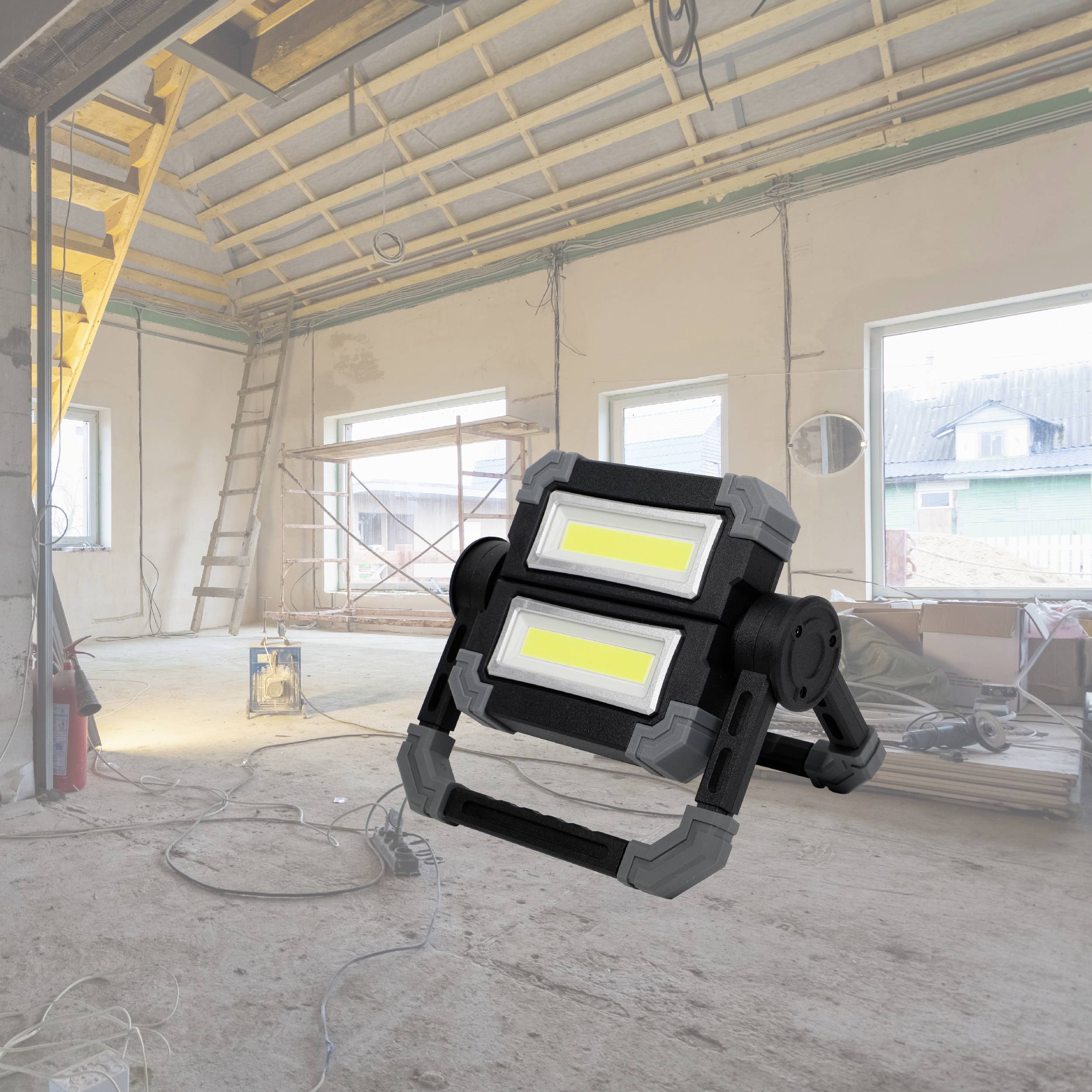 A construction site in an empty room with windows and a portable LED work light in the foreground on the floor. Ladders and building materials are visible in the background.