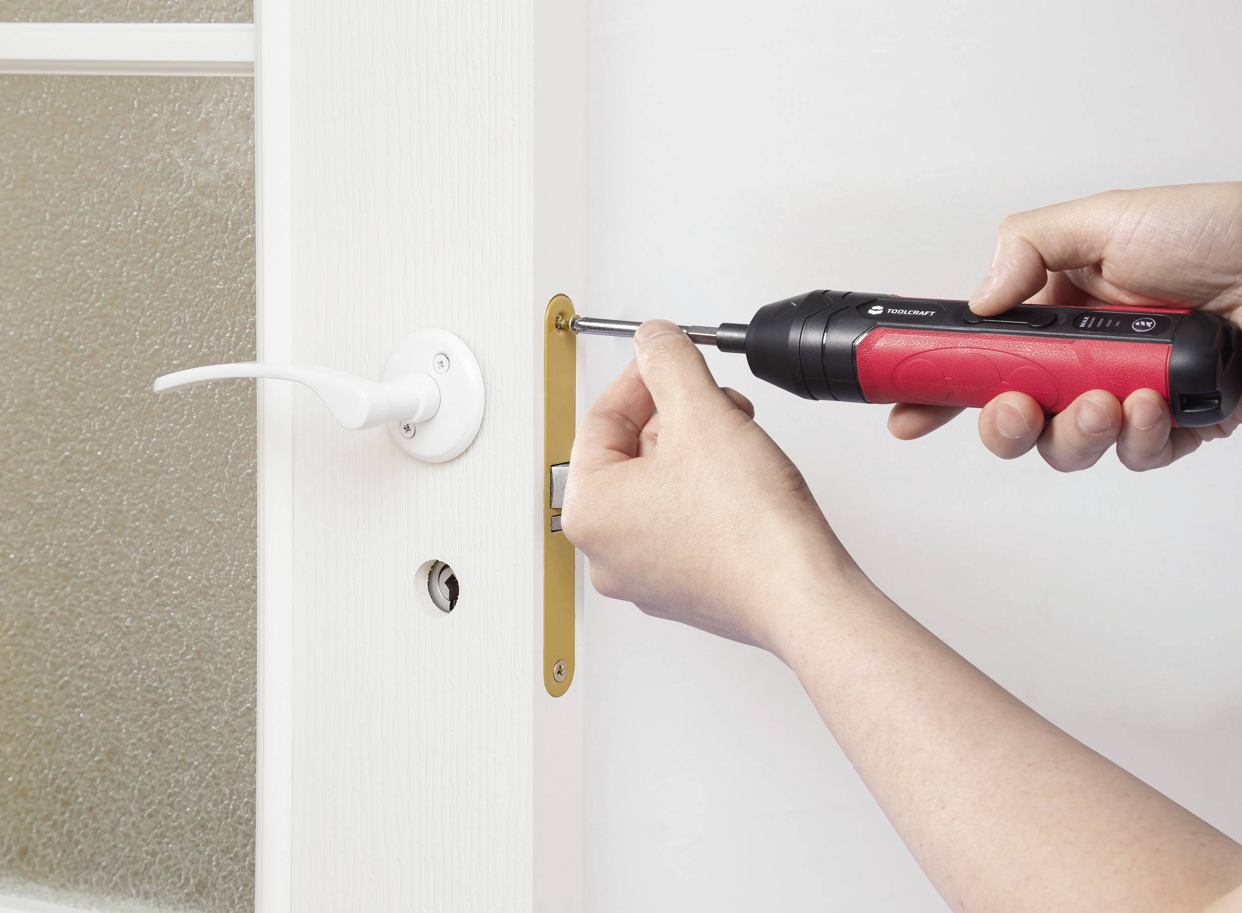 A person uses a red and black cordless screwdriver to install or remove a screw from a door handle on a white door.