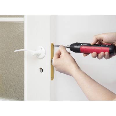 A person uses a red and black cordless screwdriver to install or remove a screw from a door handle on a white door.