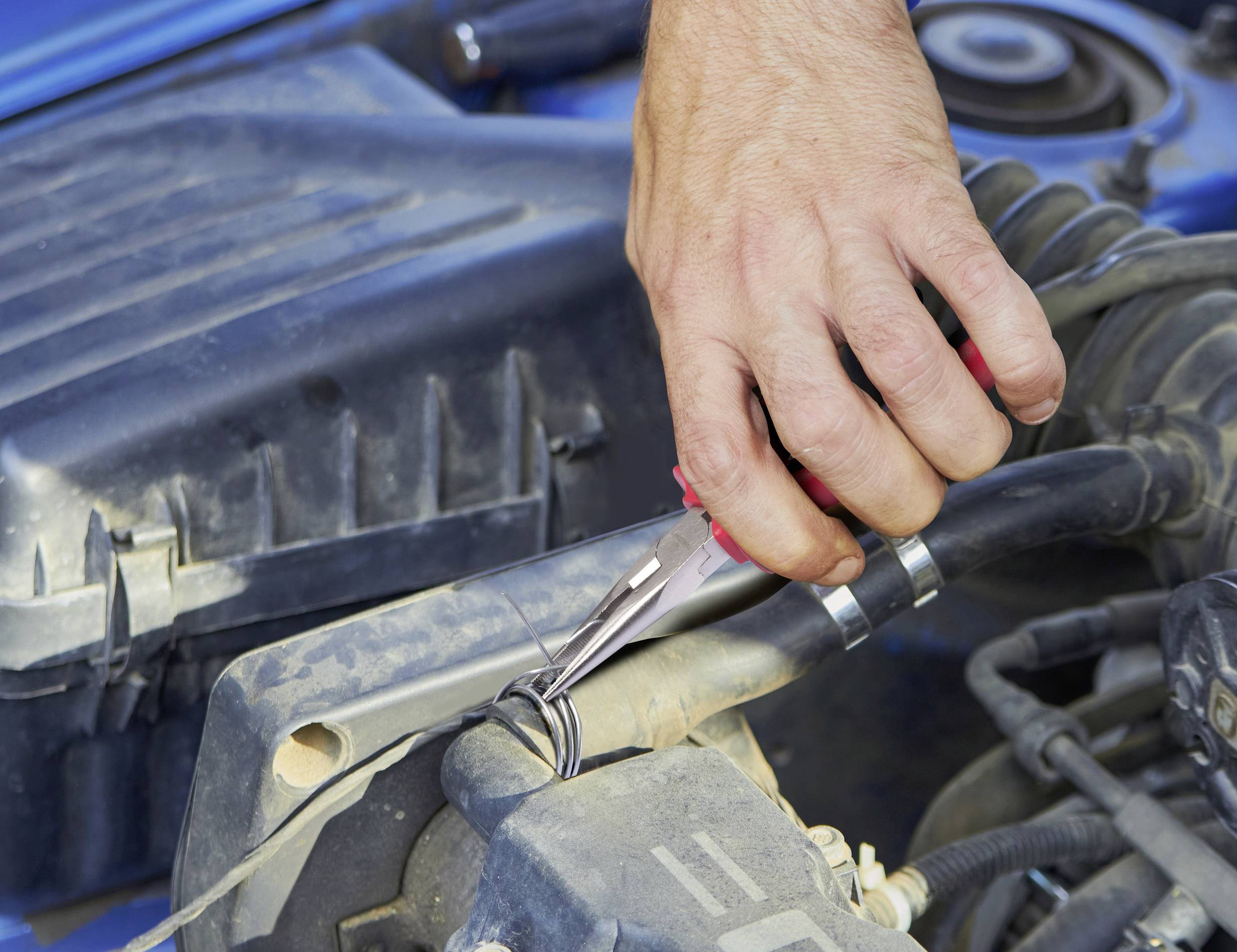 A hand uses pliers to twist wires under a car hood, performing maintenance or repairs on the vehicle's engine components.