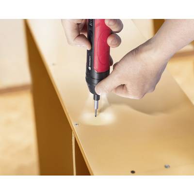 Hands using a red and black electric screwdriver to assemble furniture, driving screws into a wooden surface.
