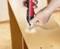 Hands using a red and black electric screwdriver to assemble furniture, driving screws into a wooden surface.