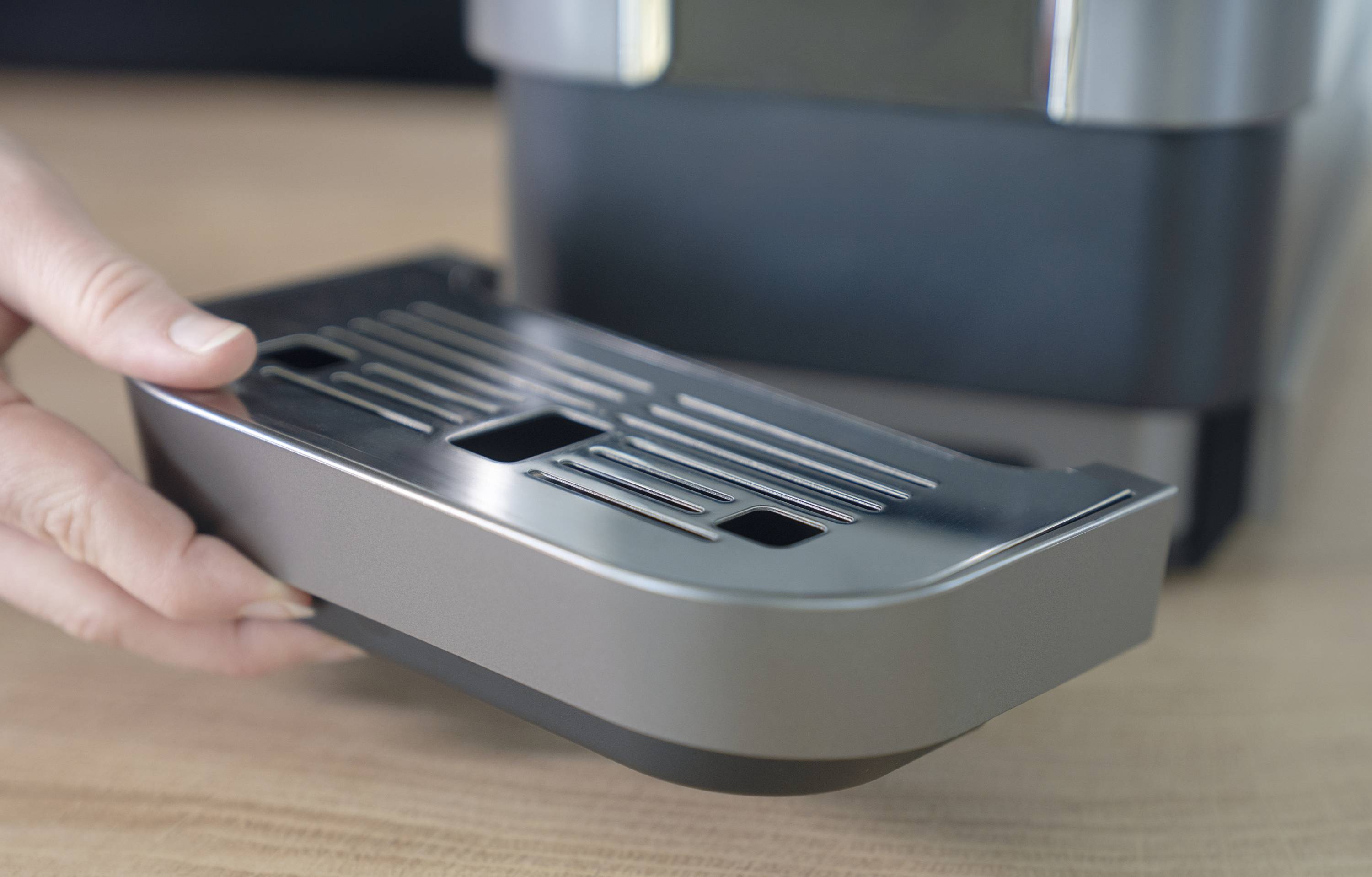 A hand removes the drip tray of a coffee machine on a wooden surface.