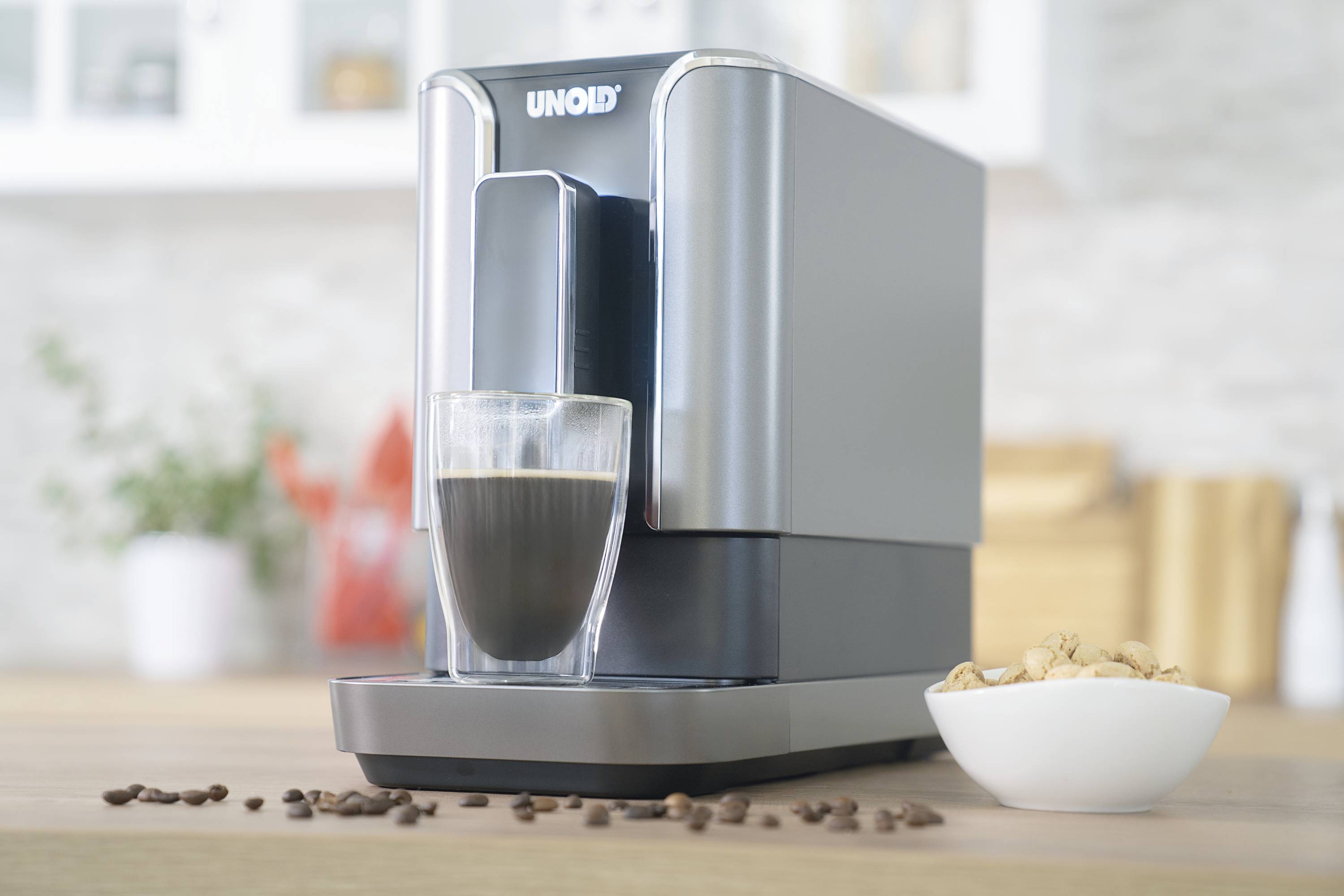 A coffee machine on a kitchen table is brewing black coffee into a glass. Beside it sits a bowl of nuts.