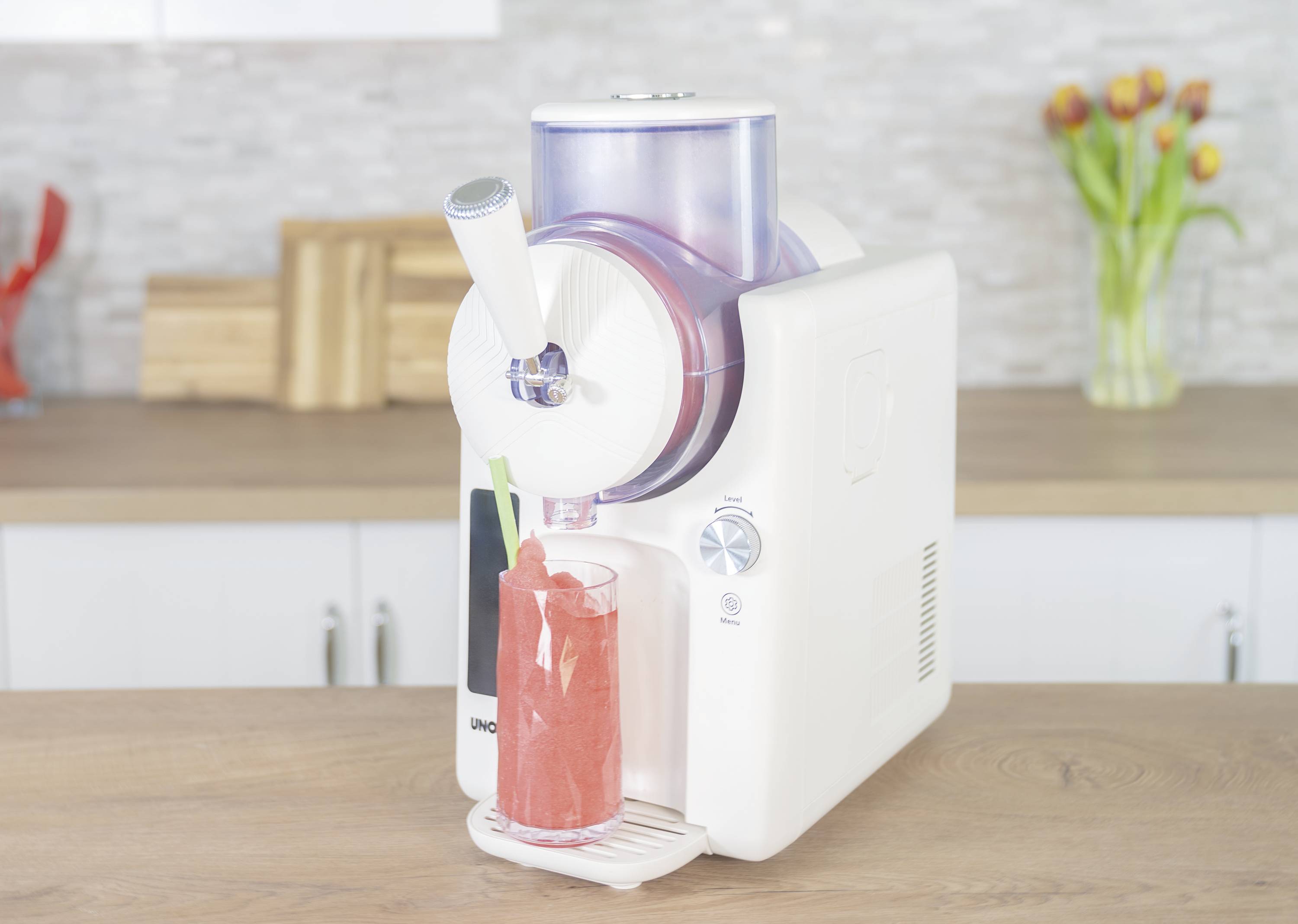 A white ice cream machine dispenser produces red sorbet that is being filled into a glass. The kitchen in the background has a modern design.