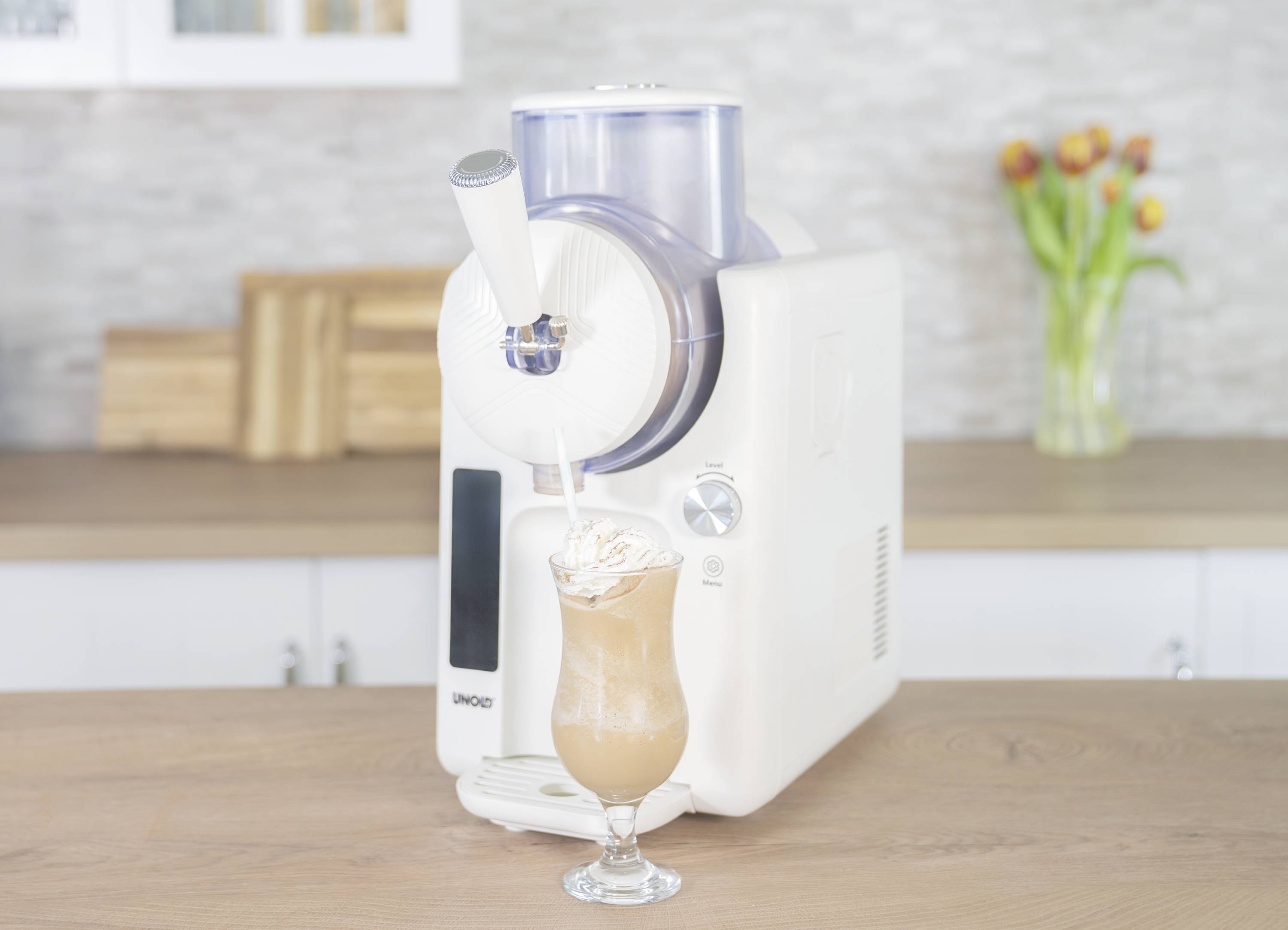 An ice cream machine fills a glass with soft serve ice cream. In the background, tulips stand in a vase on a kitchen counter.