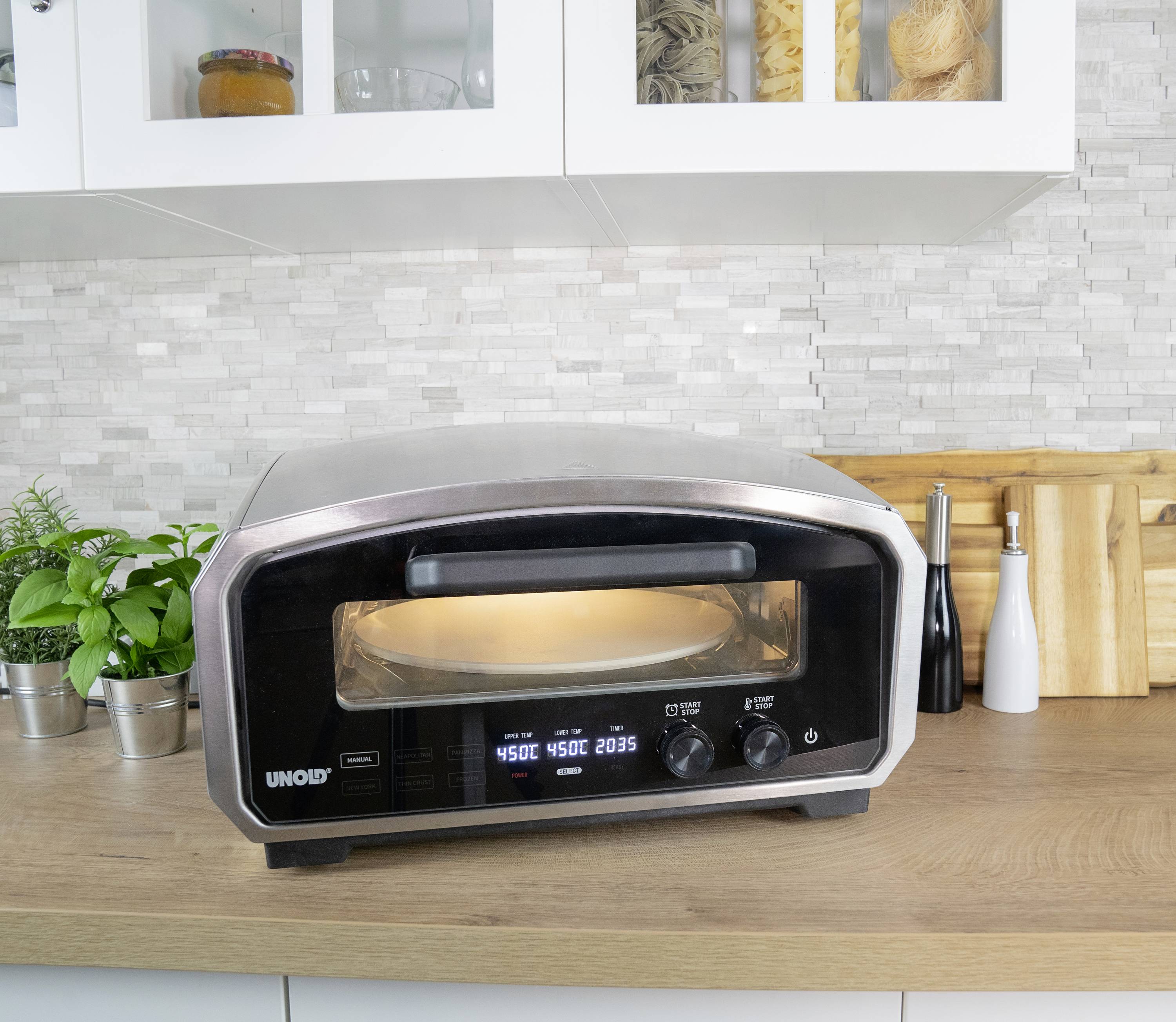 Small kitchen with an electric pizza oven on the worktop. Next to it are plants, spices, and chopping boards.