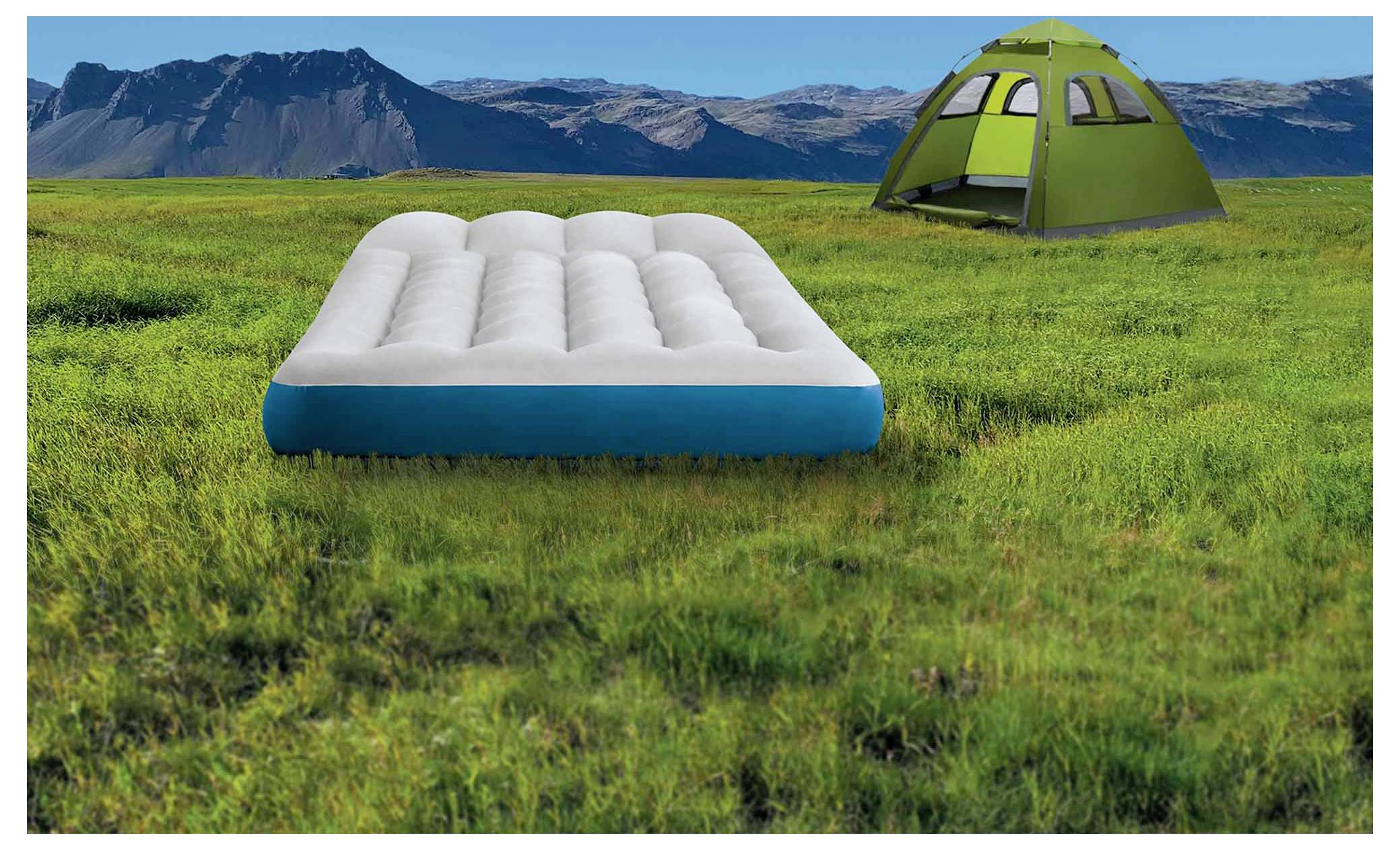 A white air mattress on green grass with a green tent in the background, set against a mountain landscape under a bright blue sky.