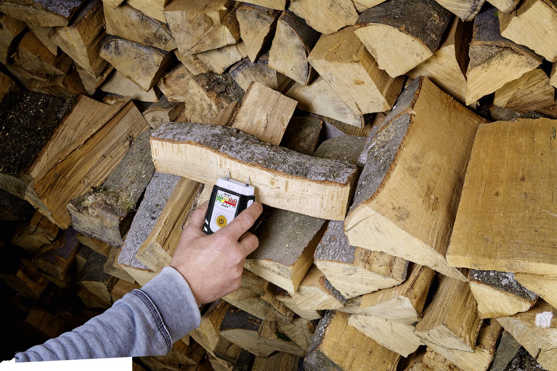 A person is measuring the moisture of a log in a pile using a wood moisture metre.
