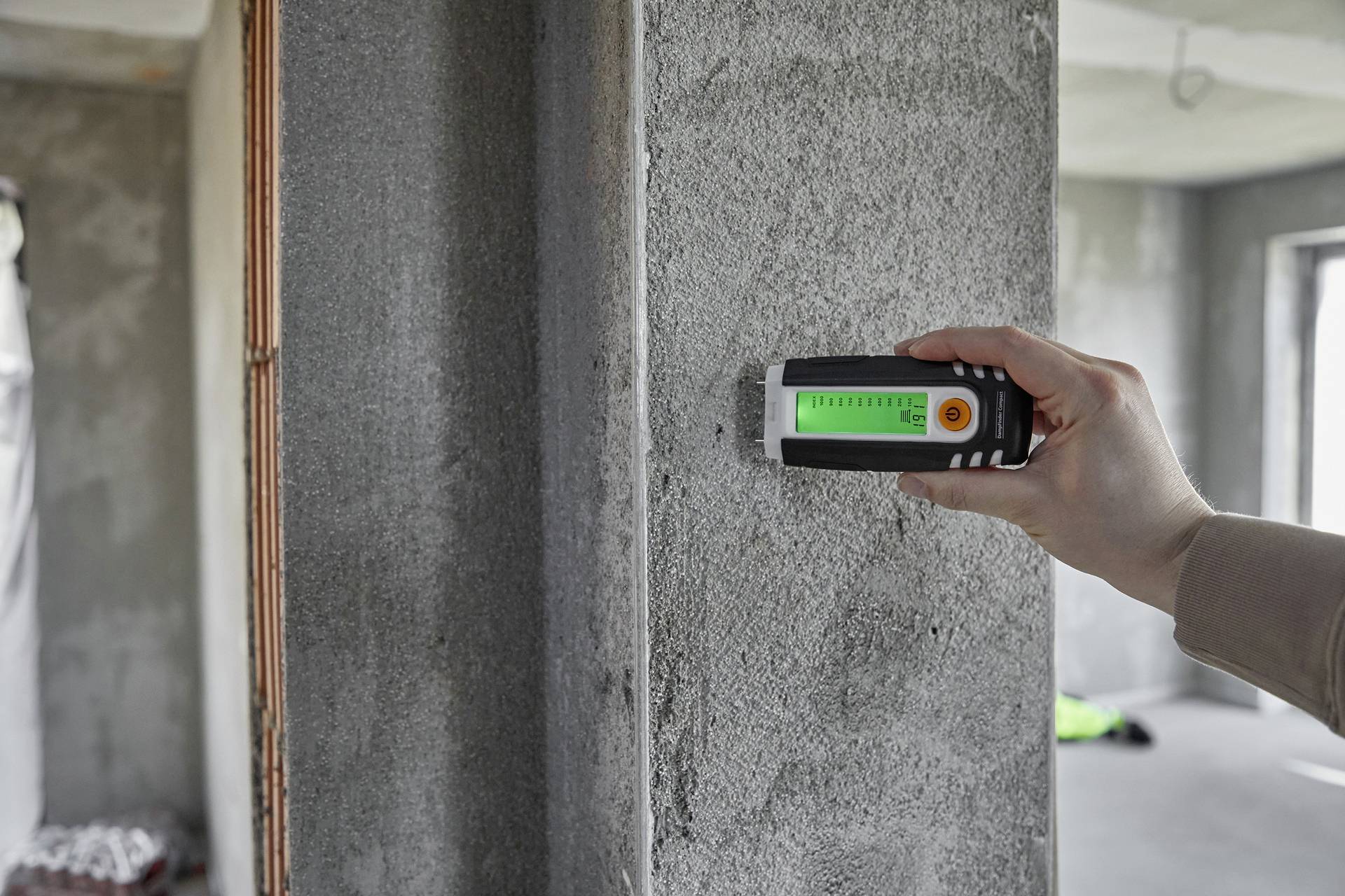 A person is measuring the moisture content in a concrete wall using a digital moisture meter. The display shows the measured reading.