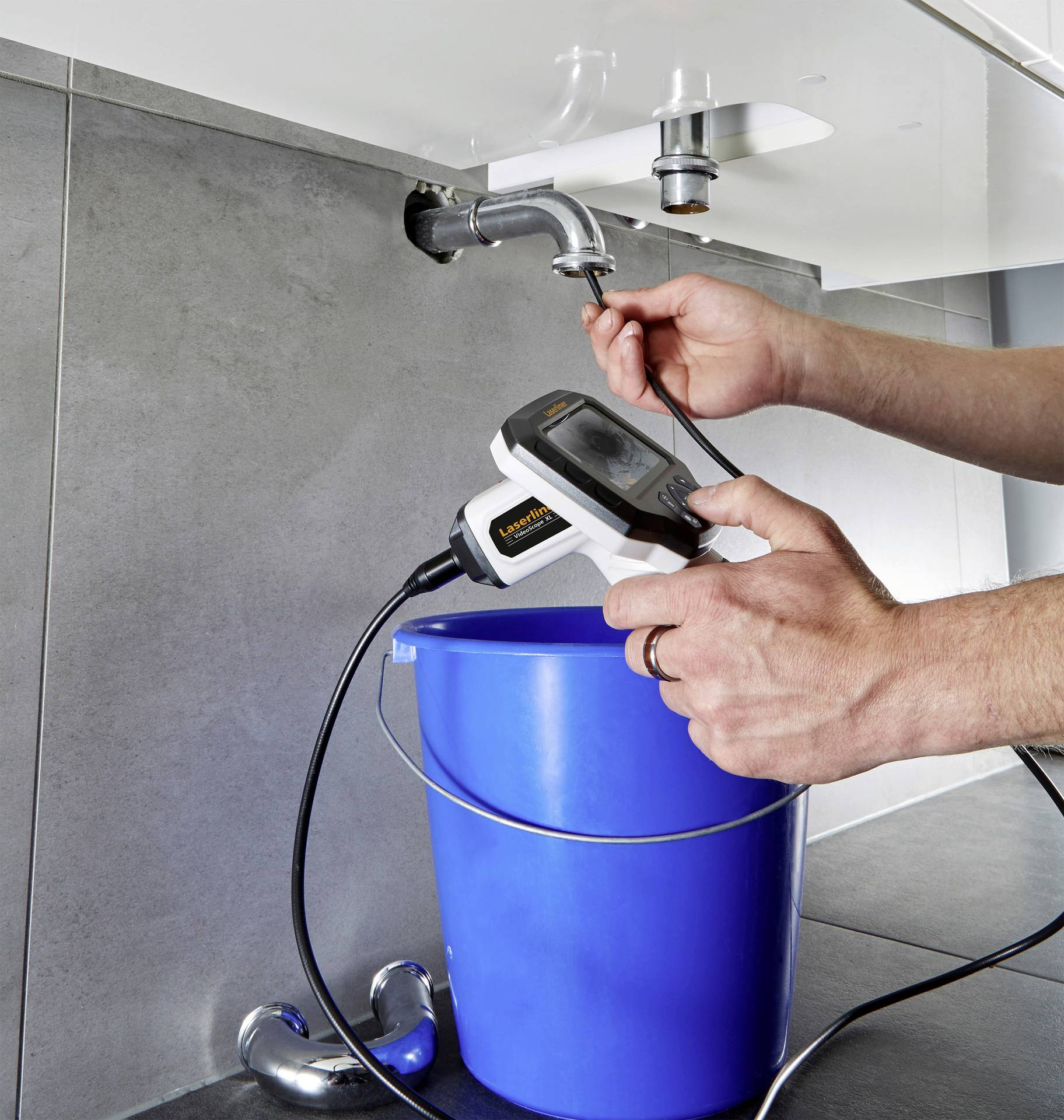 A person is using an inspection device to check the interior of a drain pipe beneath a sink. A blue bucket is positioned underneath.