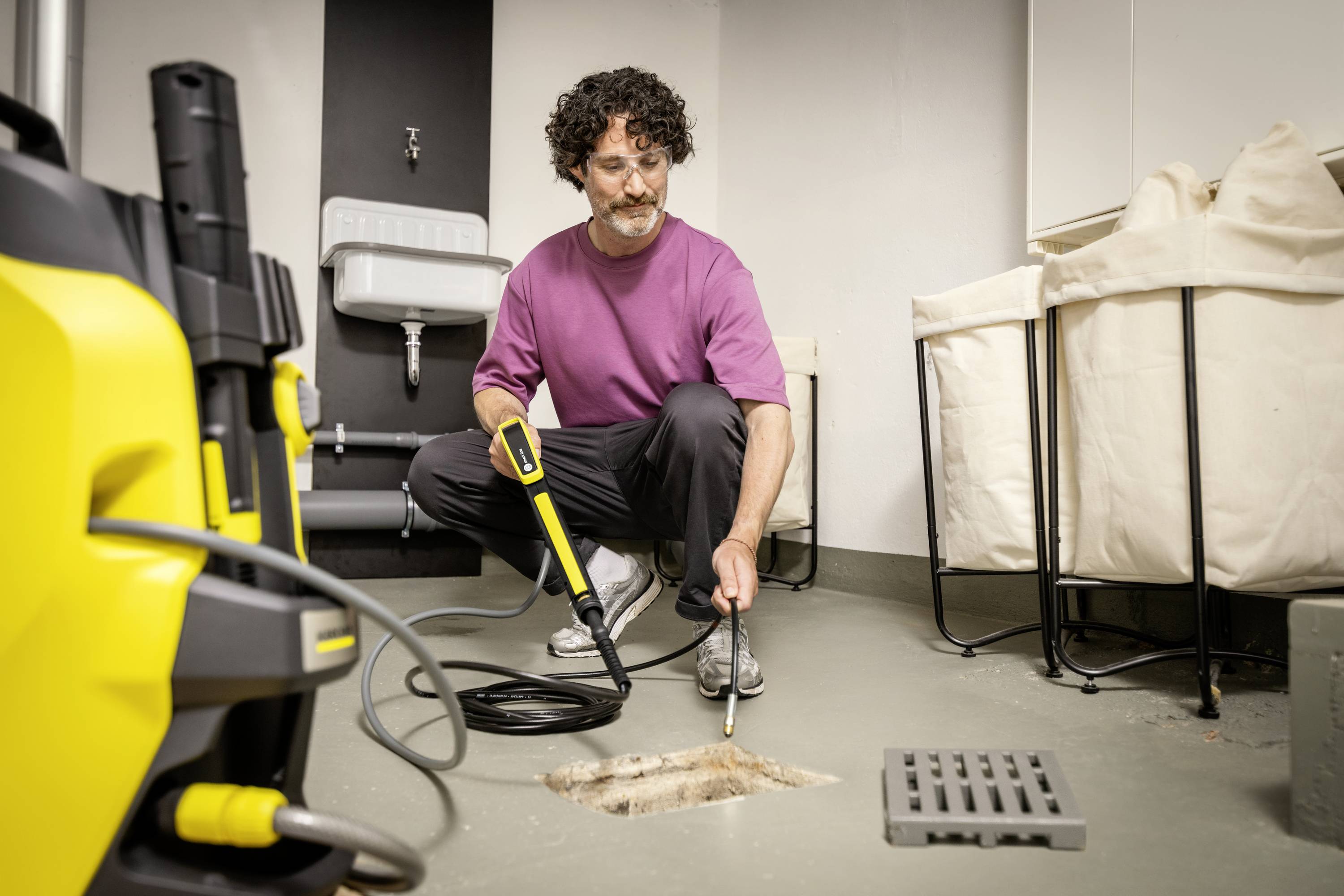 A person is using a pressure washer to clean a drain in a cellar. Sinks and baskets are visible in the background.