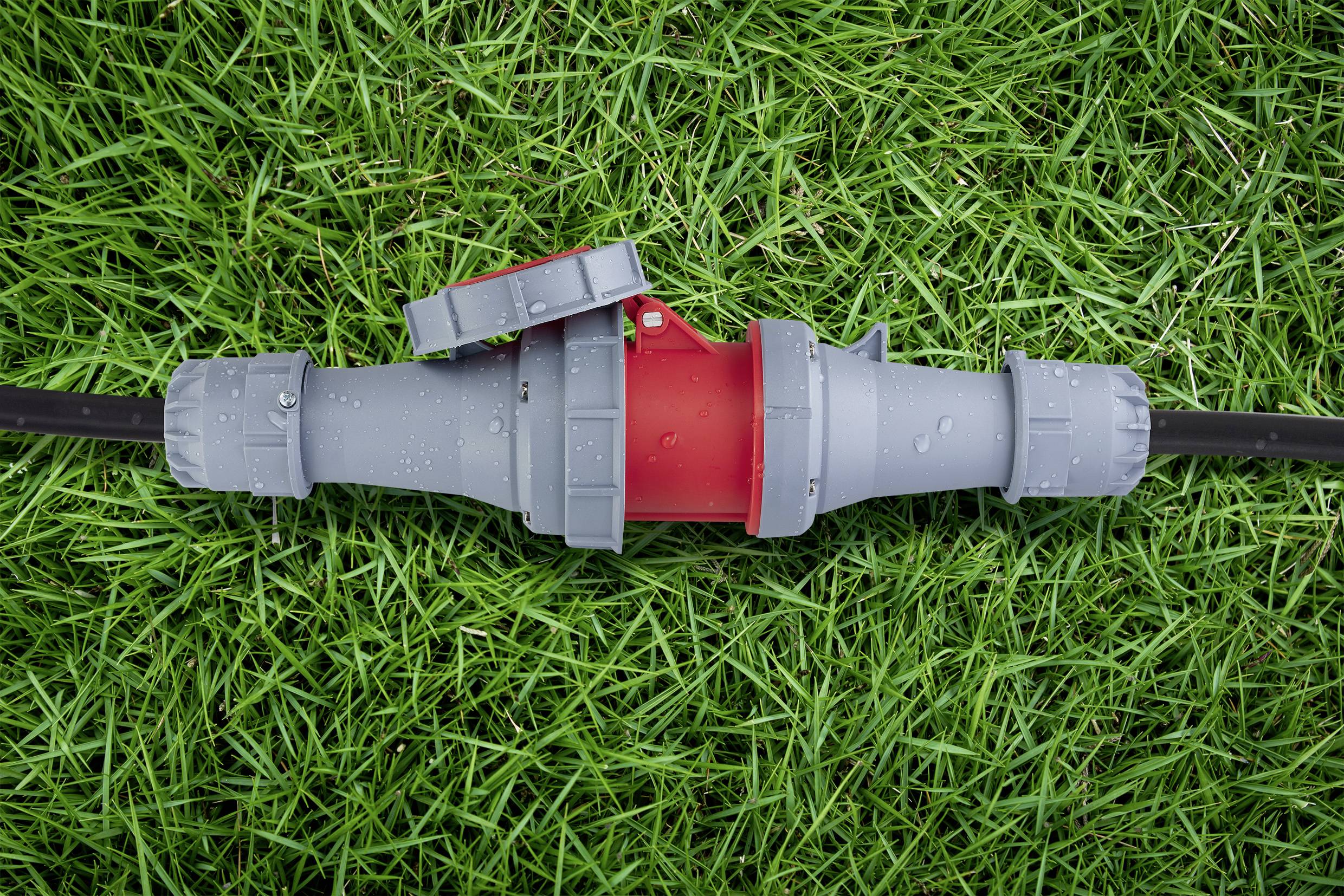 A grey and red garden hose connector lies on green grass. Droplets are visible on the surface, indicating moisture.