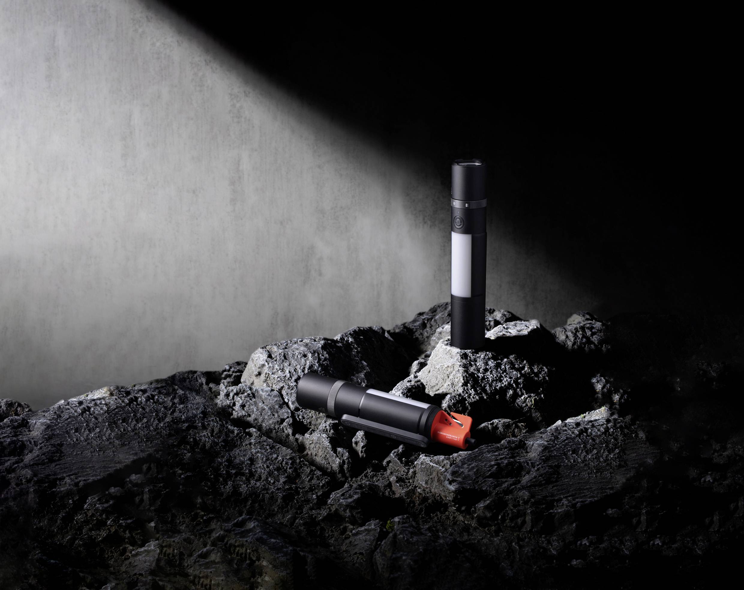 Two black cylinders on a rocky surface in the spotlight, one with a red cap. Atmosphere: mysterious and futuristic.