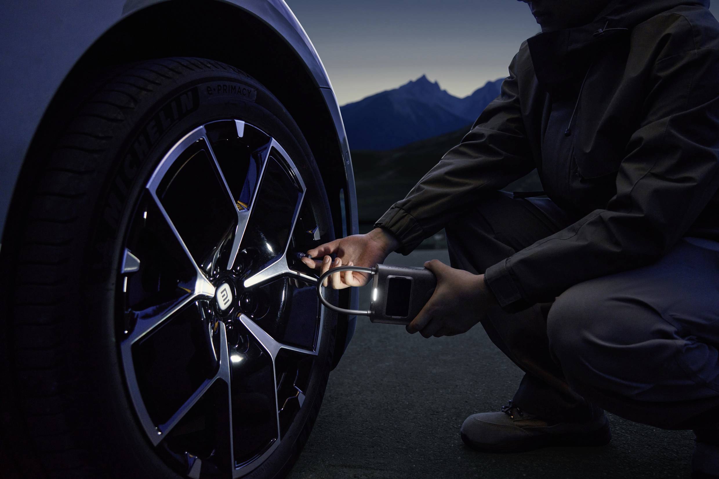 A person checks the tyre pressure of a car with a portable device during the night. Mountains can be seen in the background.