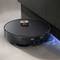 A black robotic vacuum cleaner is cleaning the floor next to a dark wooden cabinet. A blue LED is illuminating beneath the device.