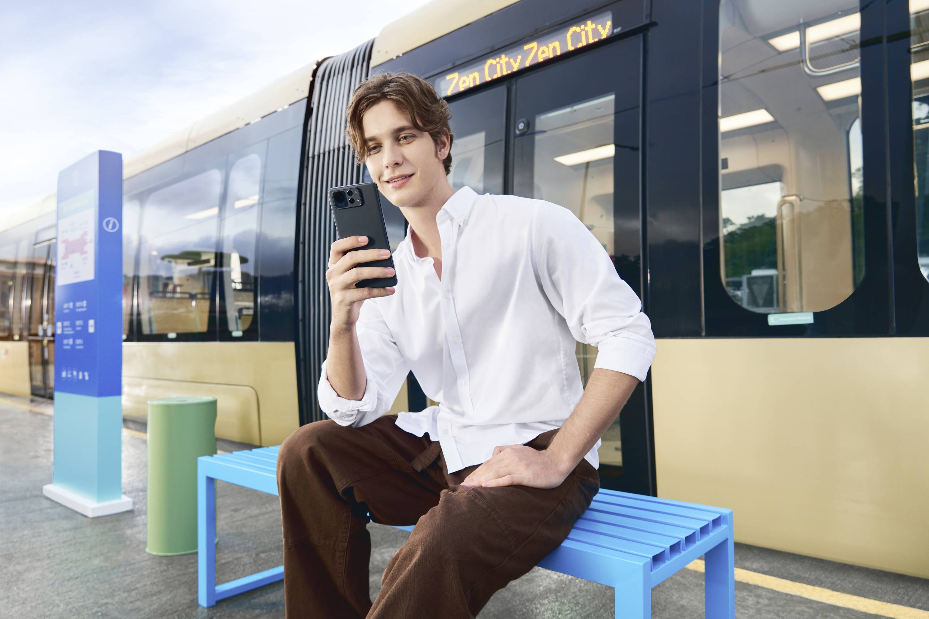 A man in a white shirt sits on a blue bench beside a railway track, looking at his smartphone. A train is in the background with a destination board reading 'Zen City'.