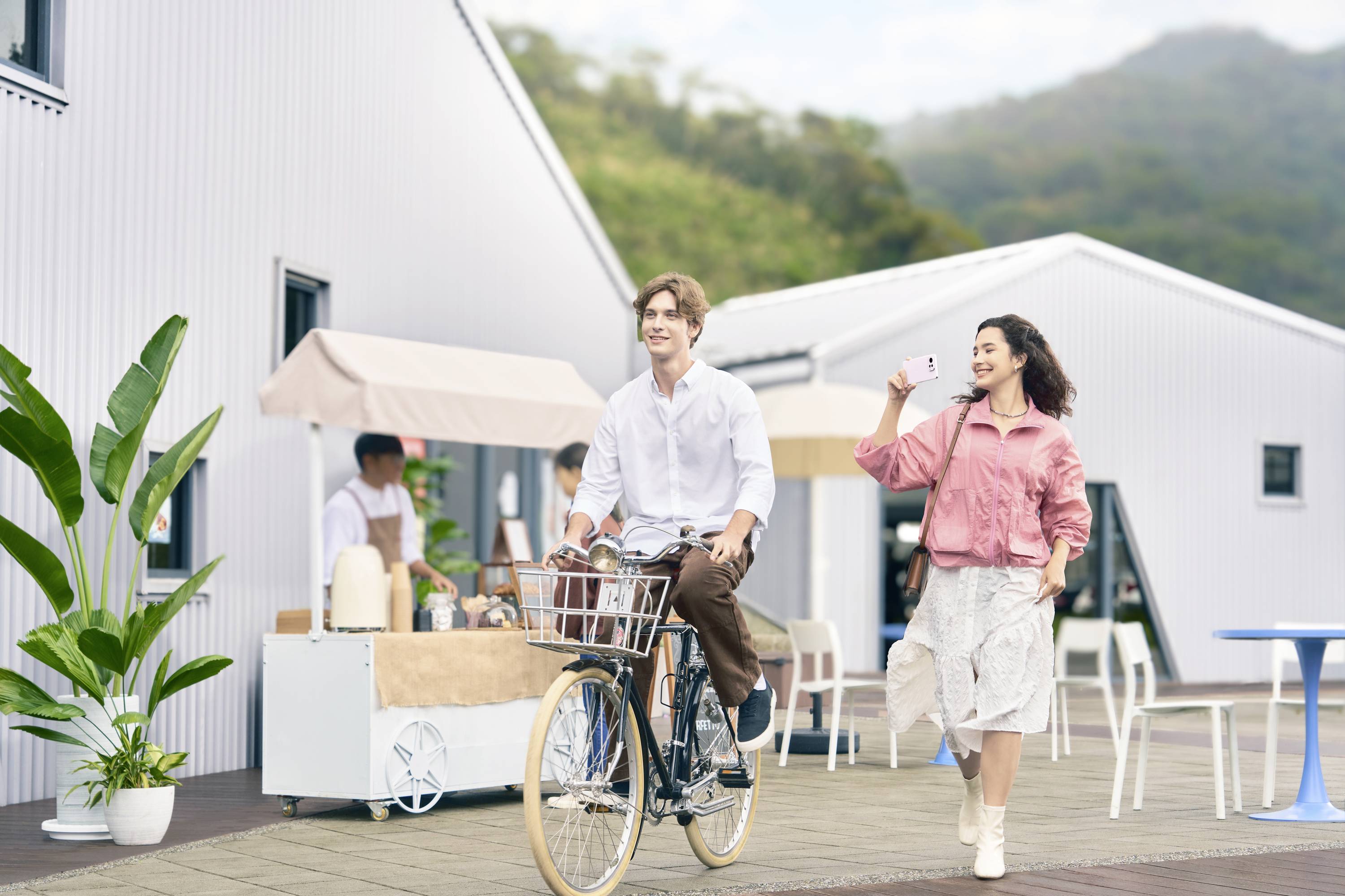 A man is happily cycling while a woman walks beside him with a drink in her hand, in front of a building with plants.