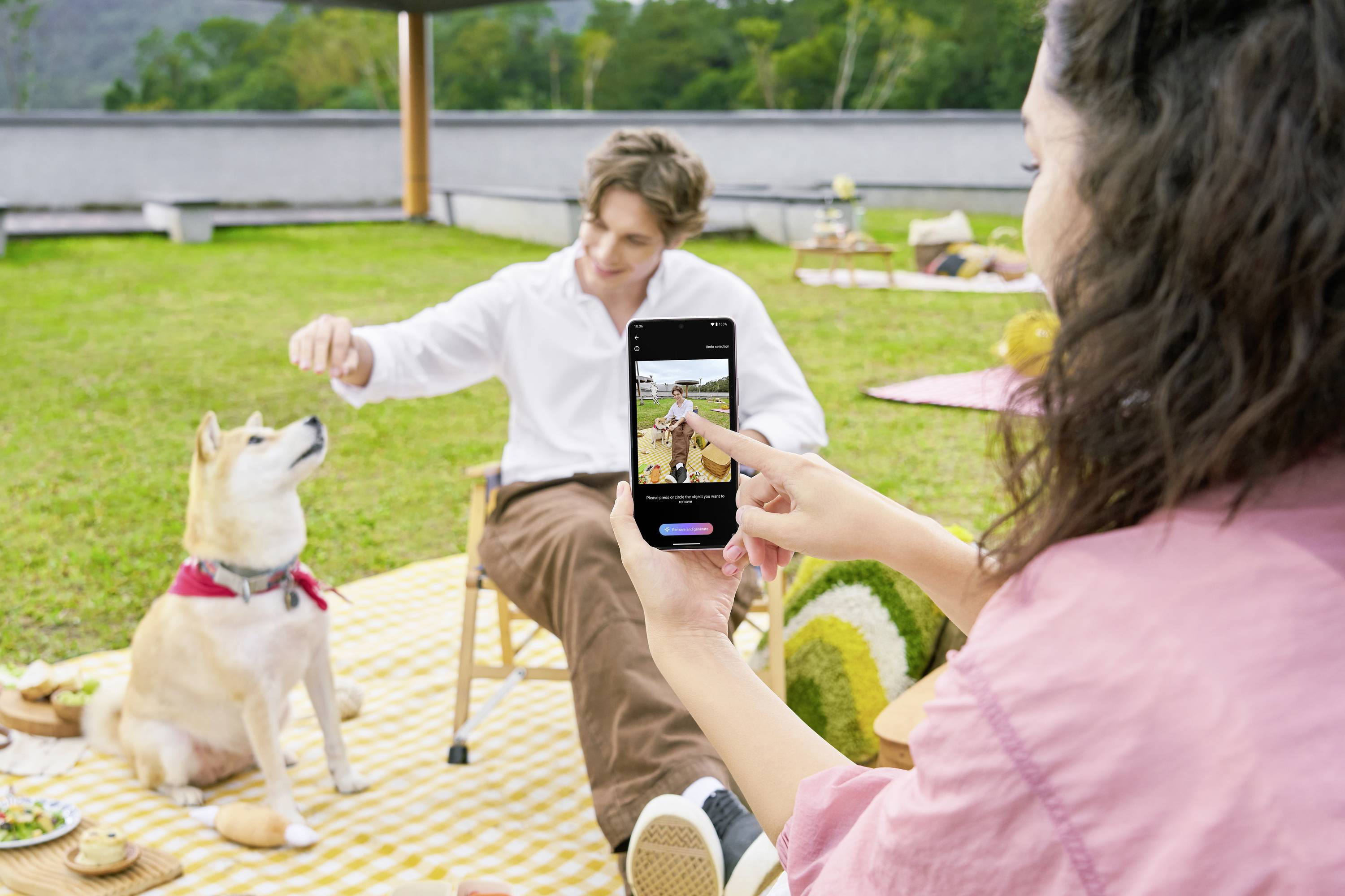A woman is taking a photo of a man giving treats to a sitting dog. Everyone is sitting on a picnic blanket outdoors.