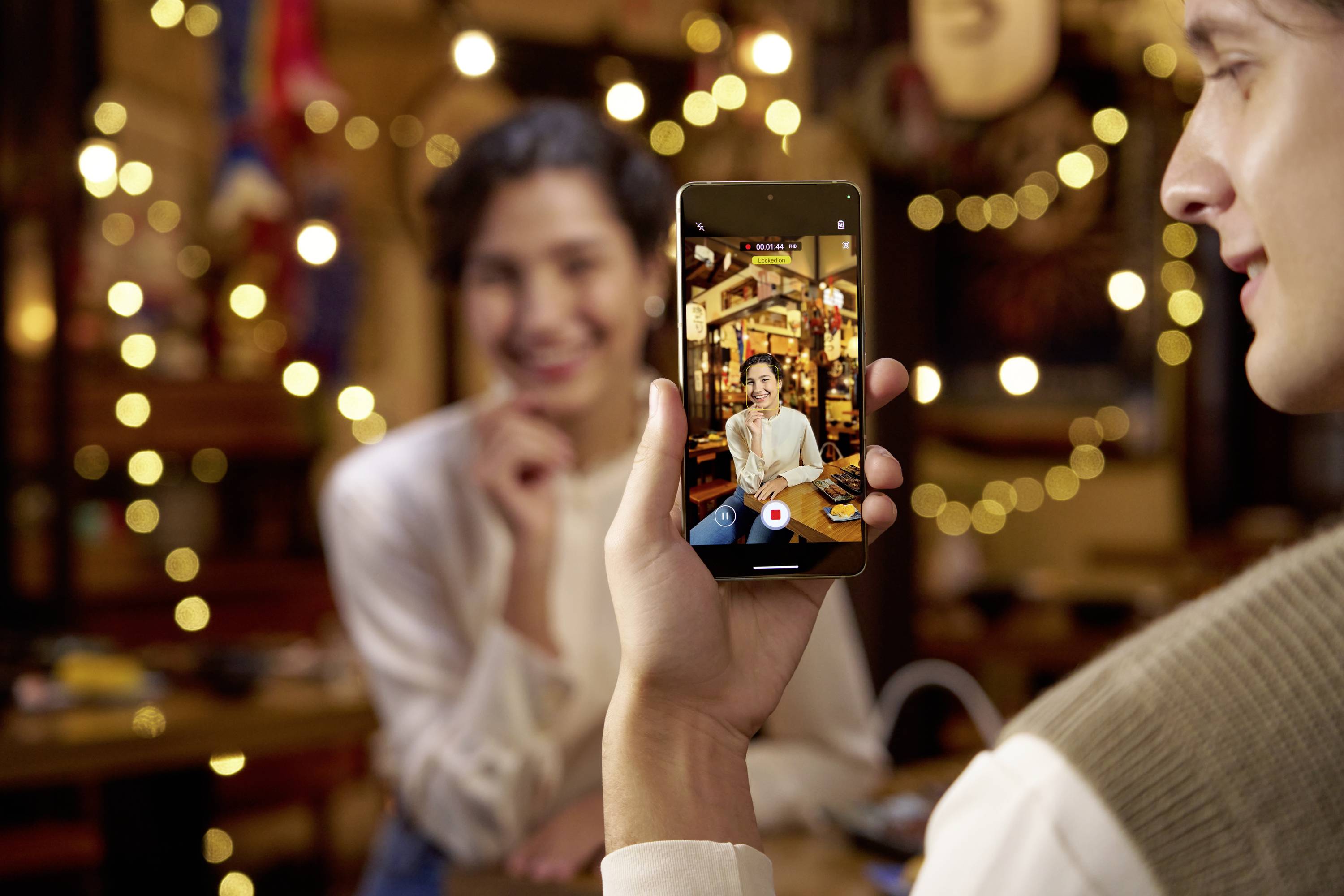 A person is holding a smartphone that shows a photo of a smiling person in the background. Lights can be seen in the background.