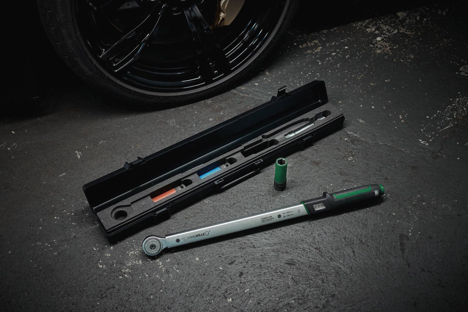 A torque wrench lies on a workshop floor next to its open case and a socket, in front of a car tyre.