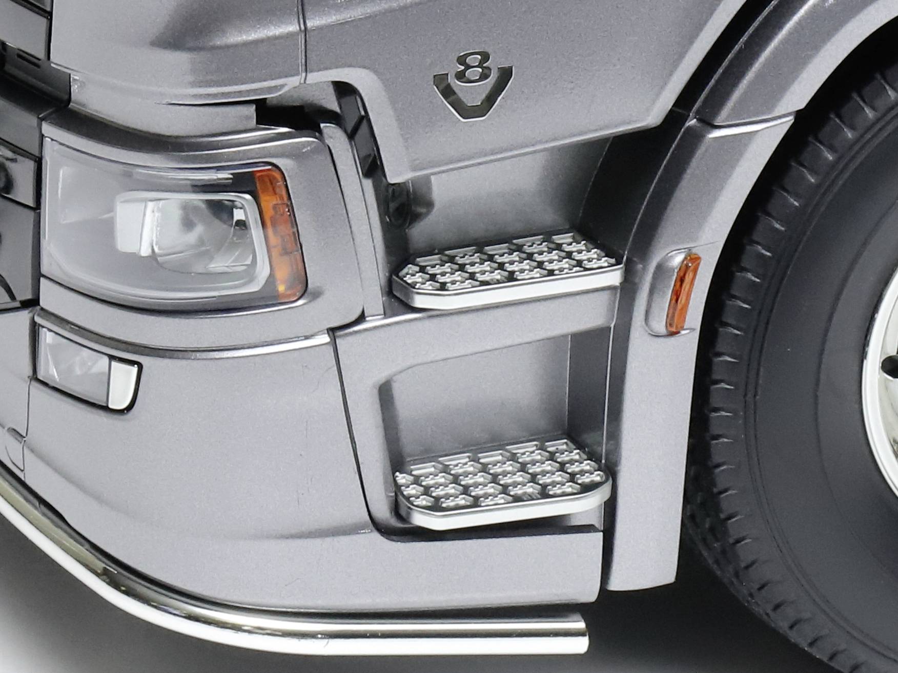 Close-up of the front section of a grey lorry with two steps, a headlight, and a tyre.