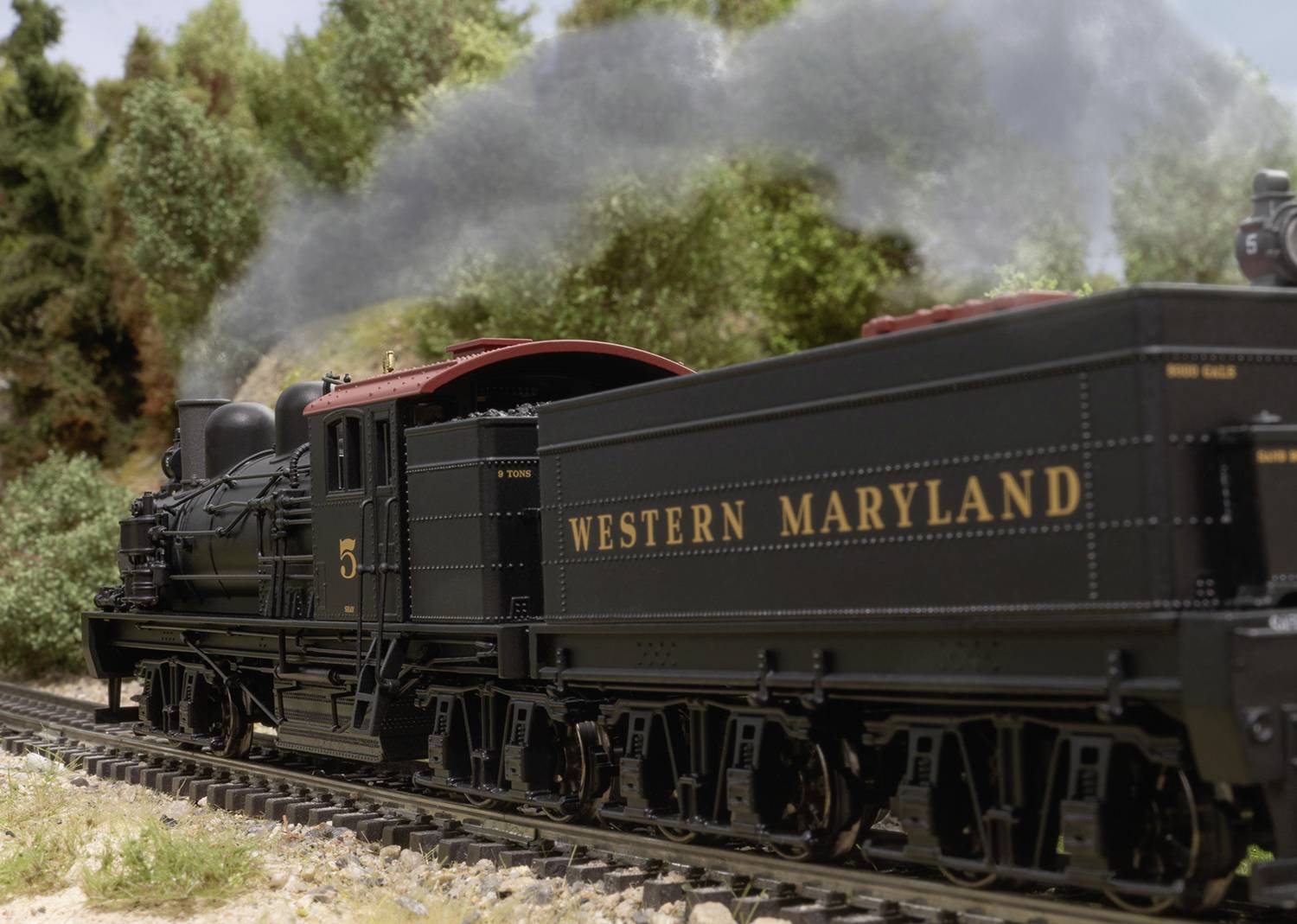 A steam locomotive of the 'Western Maryland Railroad' steams through a green, wooded landscape on railway tracks. Historic train representation.