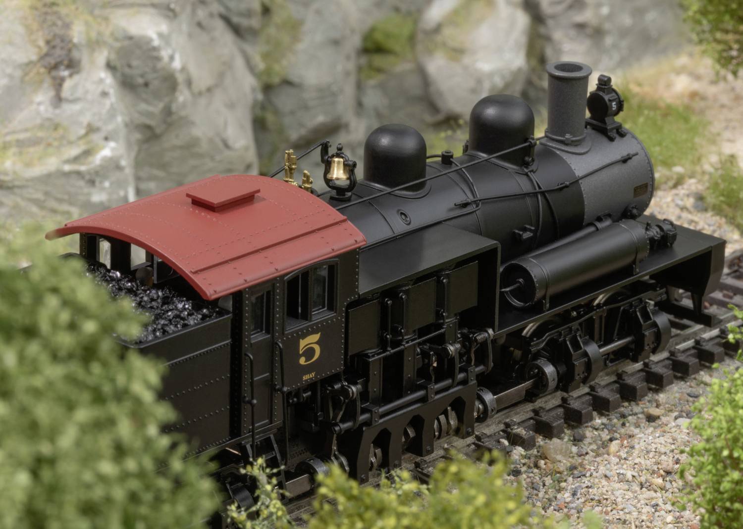 Black steam locomotive with a red roof and the number '5' on the track, surrounded by rocks and grasses in a model railway scene.