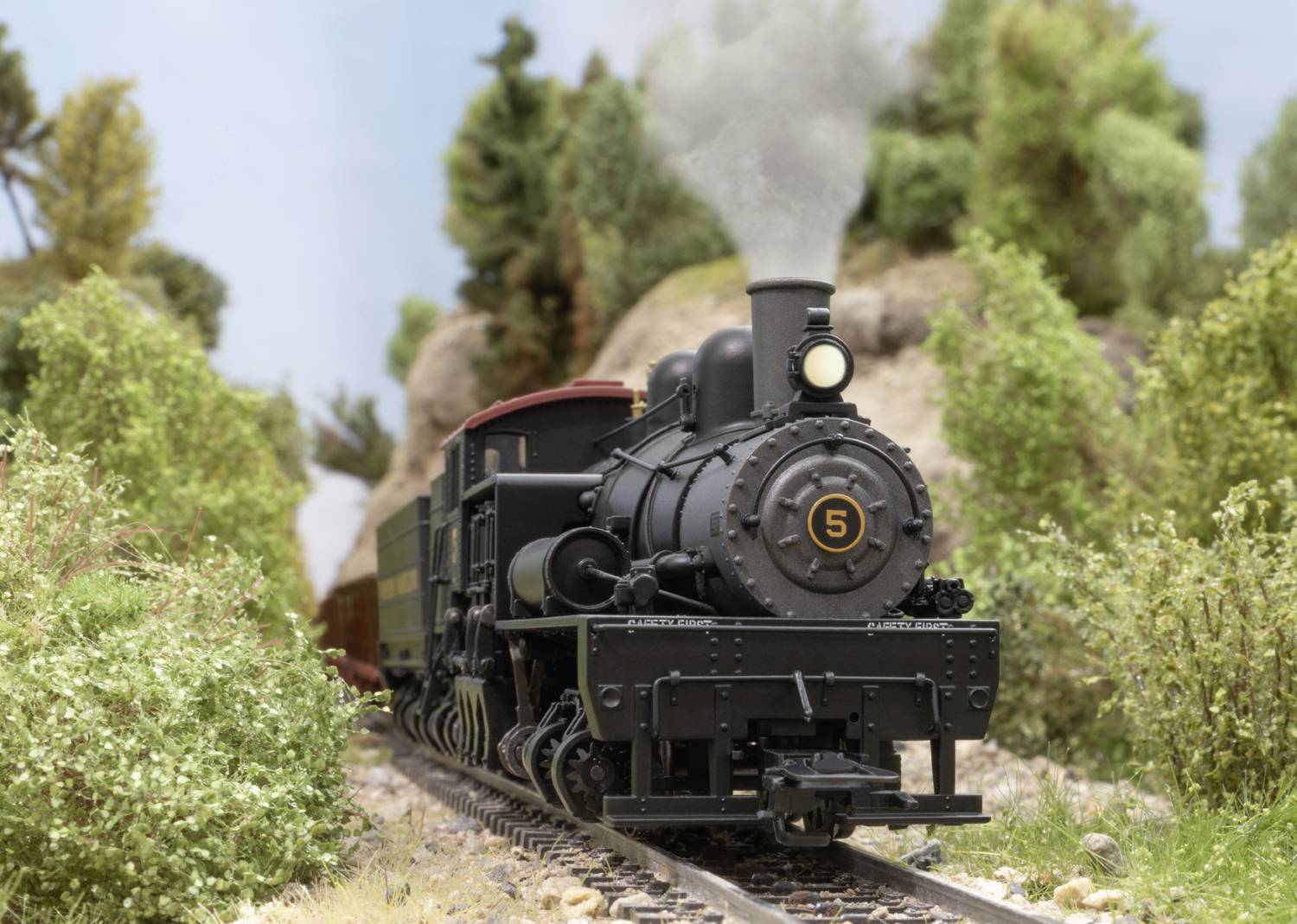 A black steam locomotive with the number '5' is travelling through a green, wooded landscape. Smoke rises from its chimney.