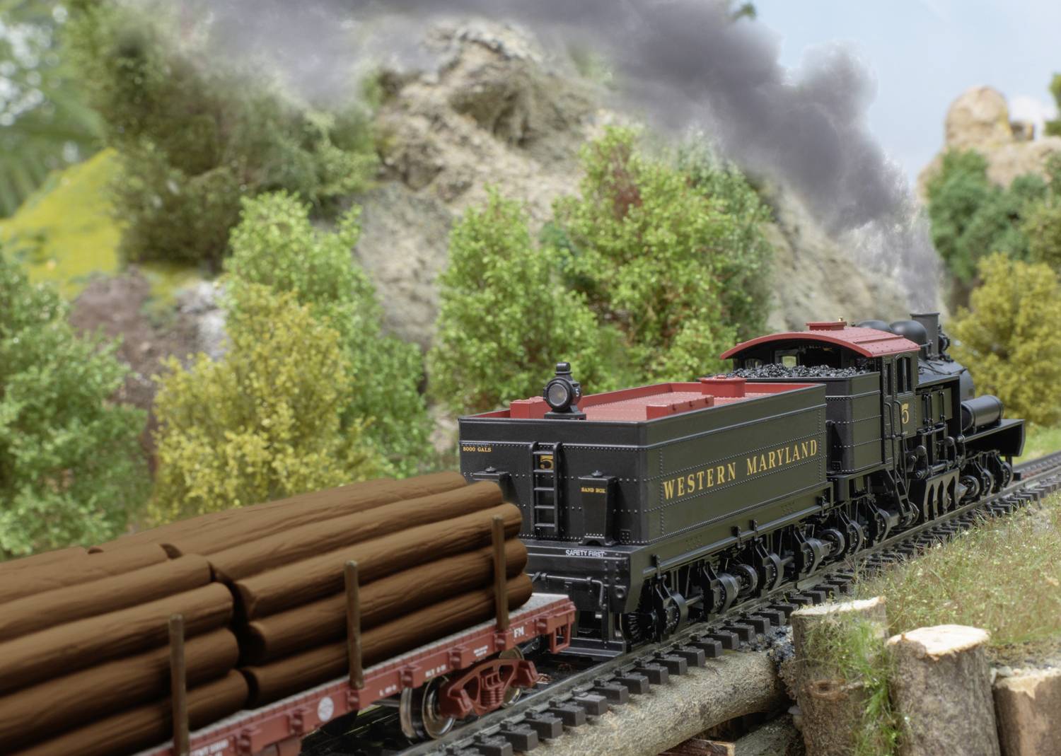 A black and red model train from 'Western Maryland' is transporting logs through a miniature landscape with trees and hills.