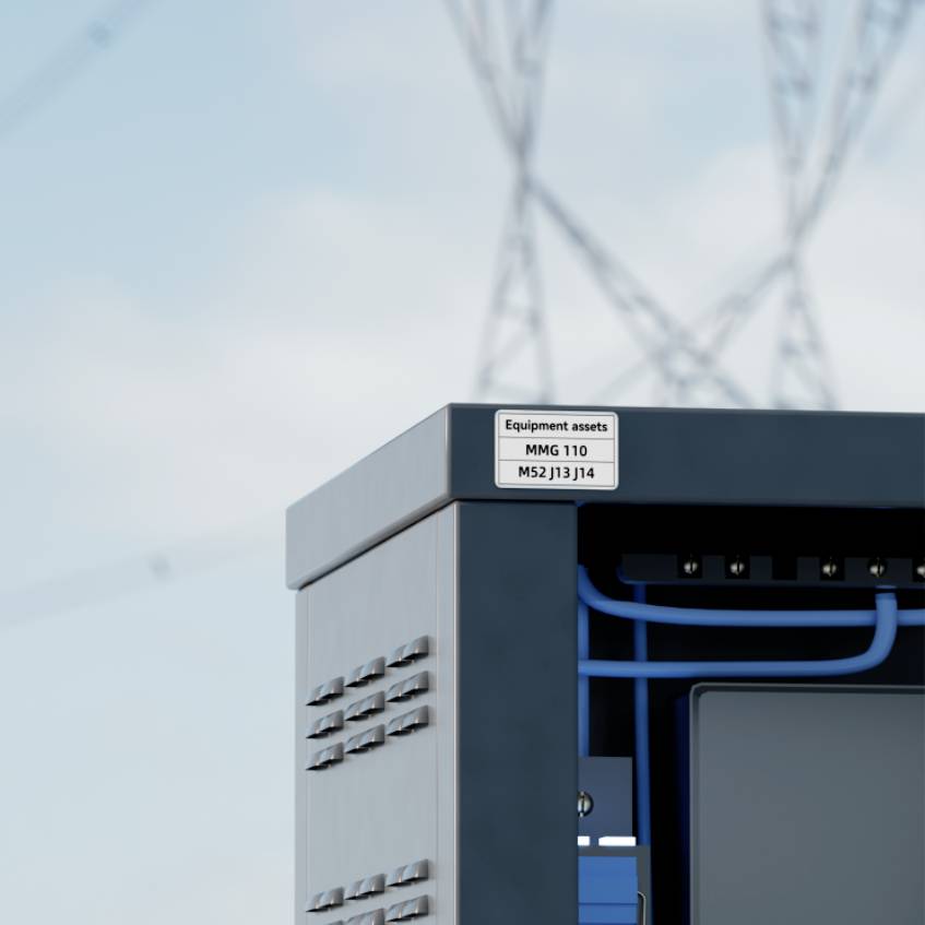 Electrical cabinet outdoors beneath electricity pylon, sticker reading 'Equipment assets MMG 110 M52 J13 J14' visible.
