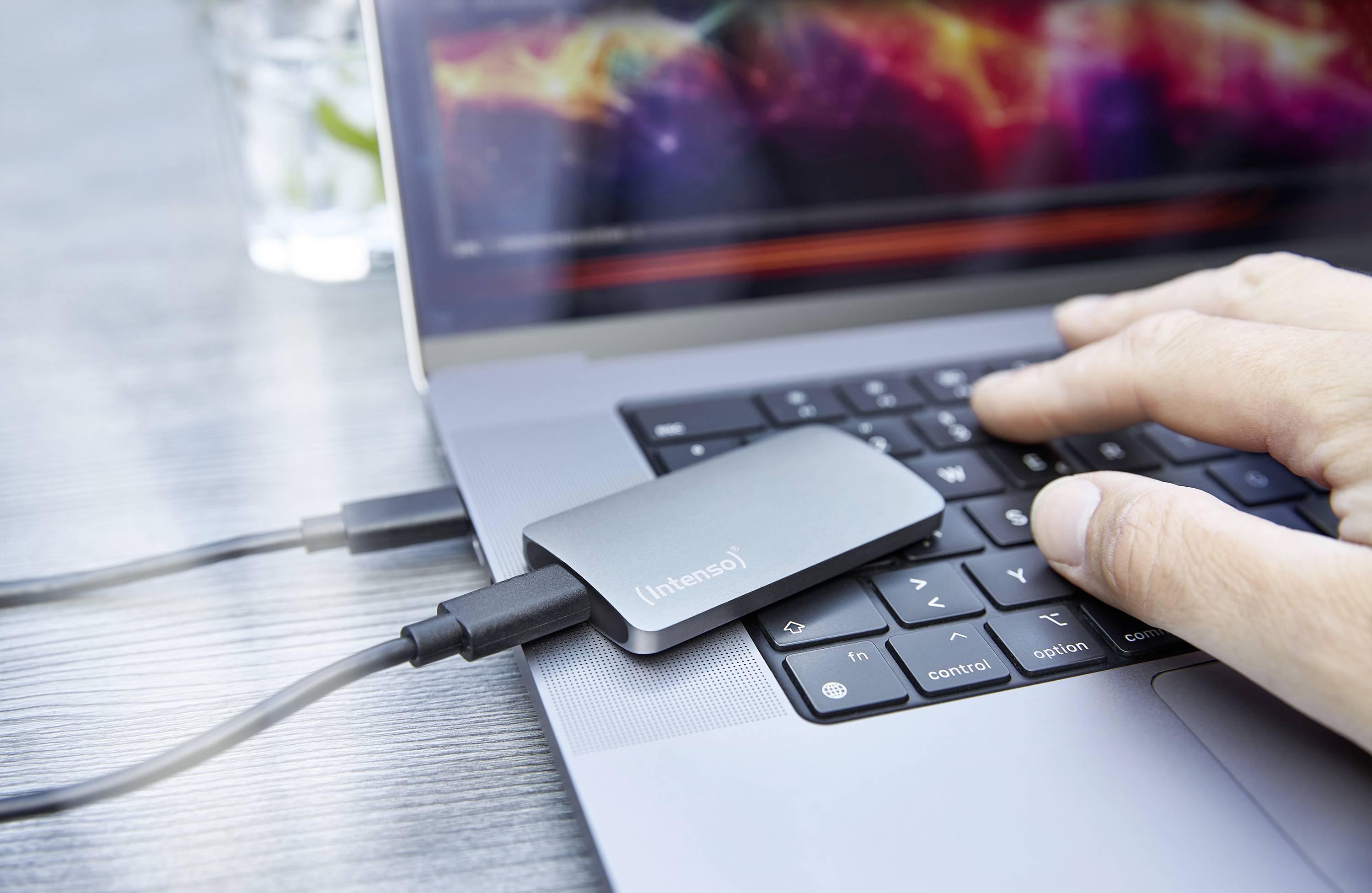 A hand is typing on a laptop keyboard, with an external hard drive connected. The screen displays colourful patterns.