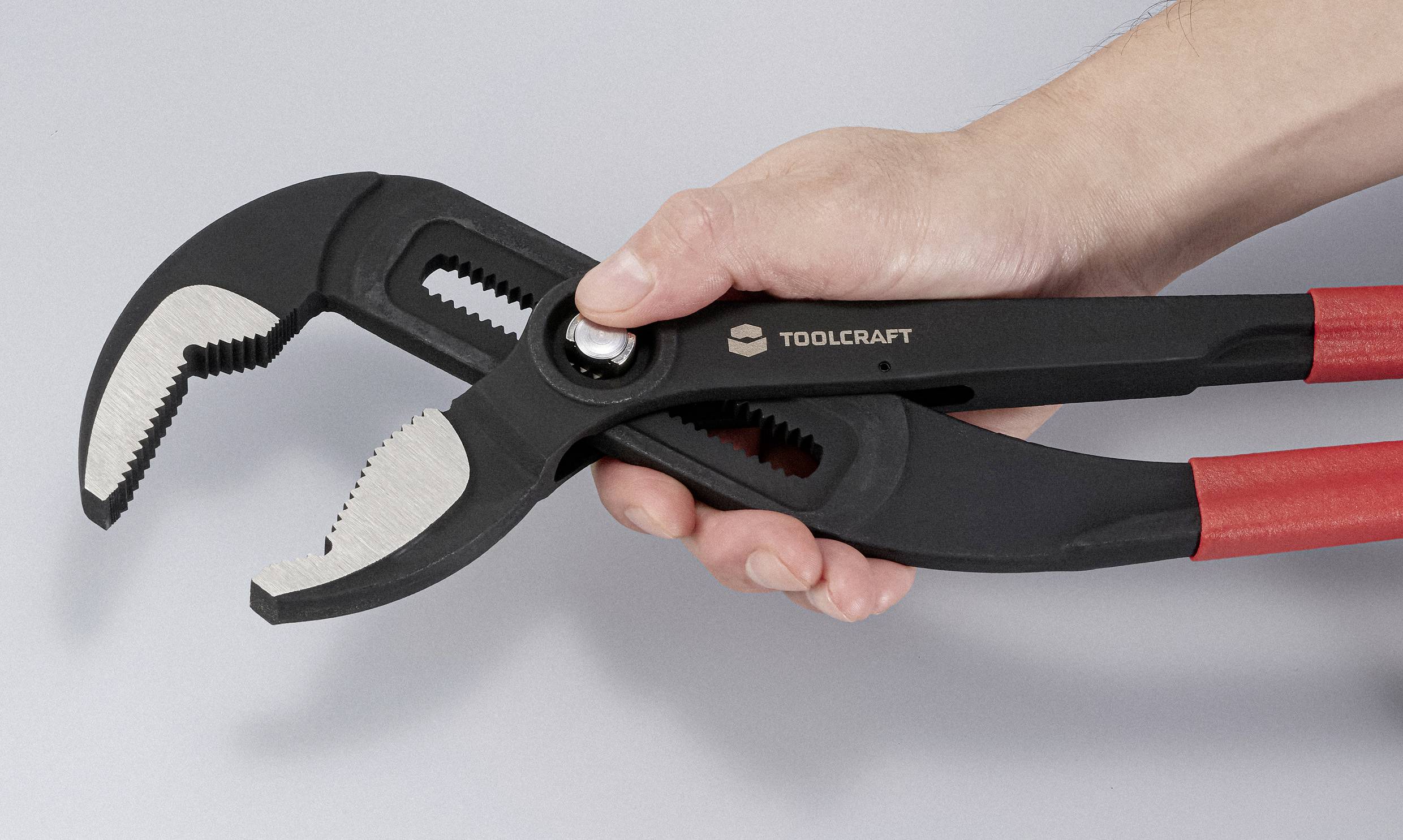 A person holds a pair of adjustable pliers with red handles, showing its open jaws and serrated gripping surfaces, against a light gray background.