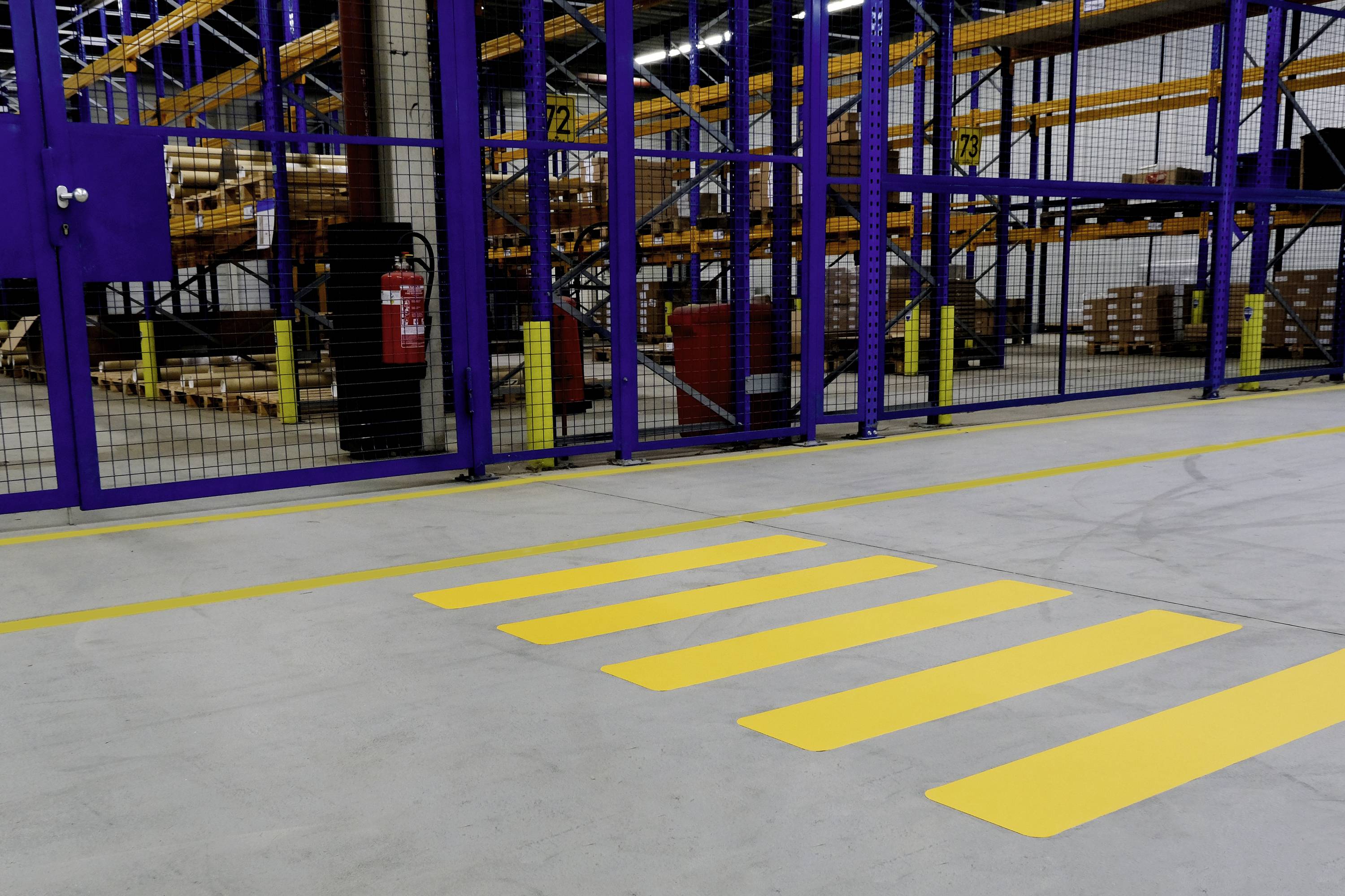 Yellow zebra crossing in a warehouse in front of a metal fence, with a fire extinguisher nearby. In the background, numerous shelves stacked with boxes.