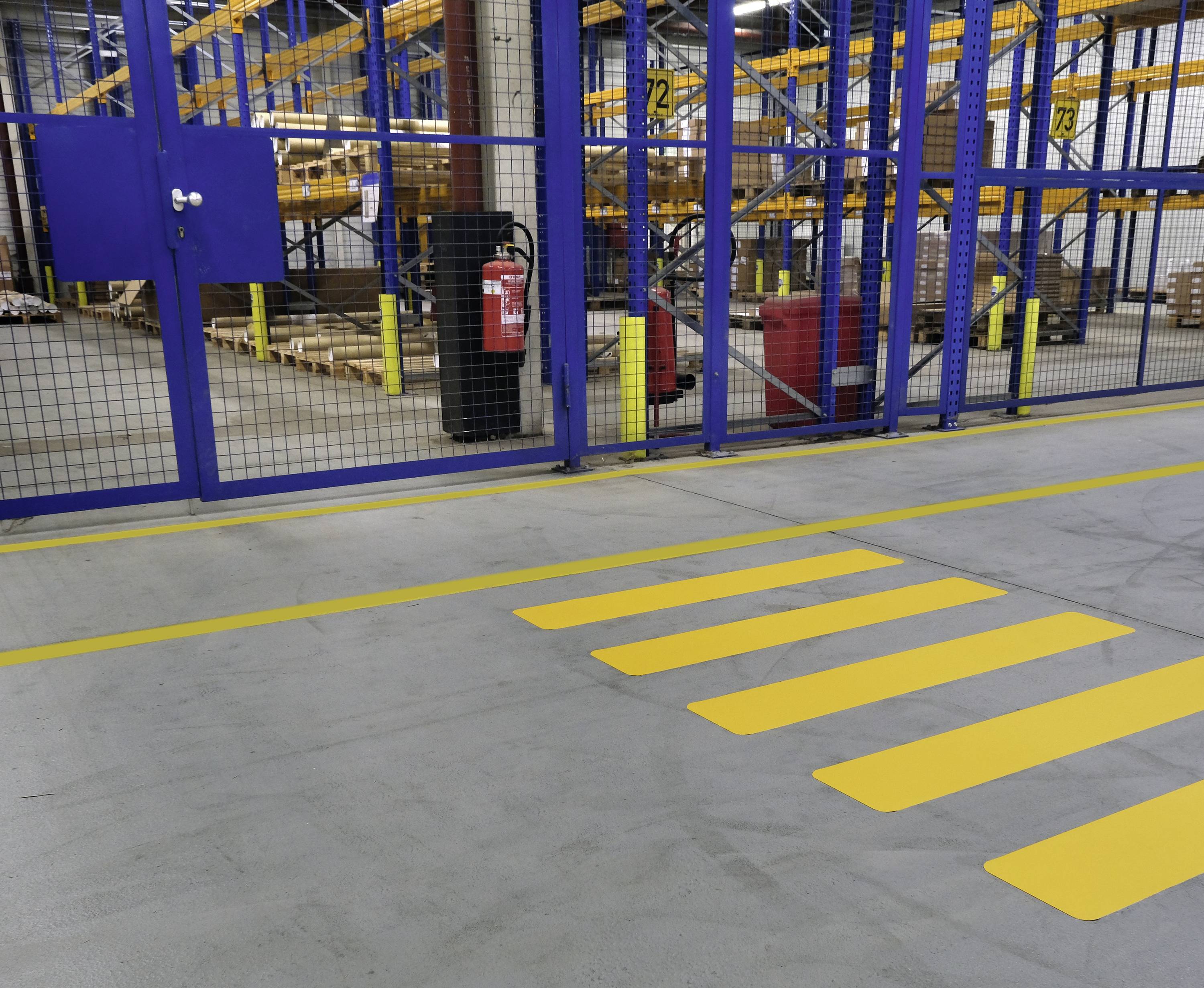 Yellow zebra crossing on grey floor in a warehouse, leading to a mesh gate. Shelves and warehouse equipment are visible in the background.