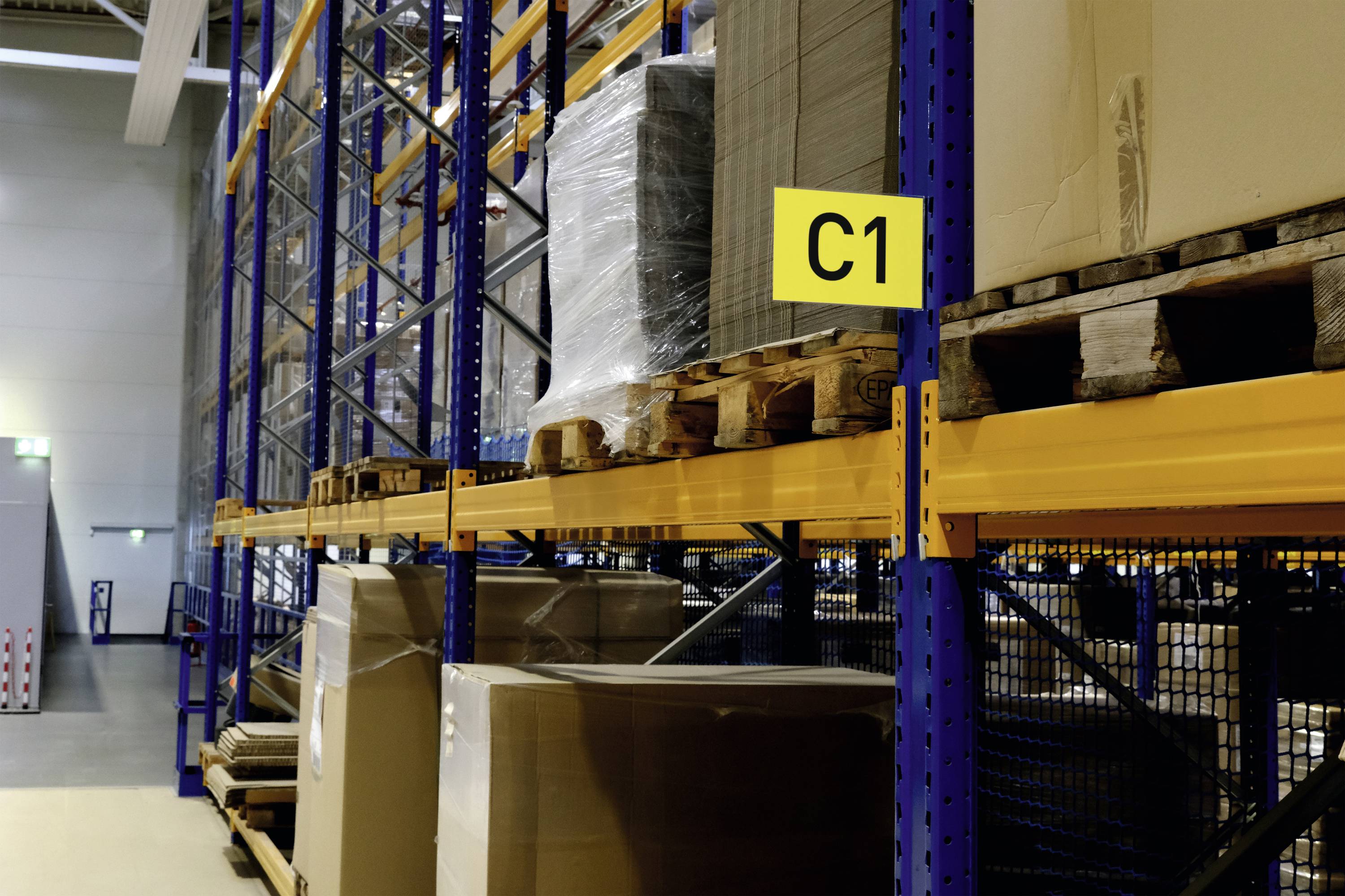 A storage rack with a yellow 'C1' sign, filled with large pallets wrapped in plastic sheeting, in a well-lit warehouse.