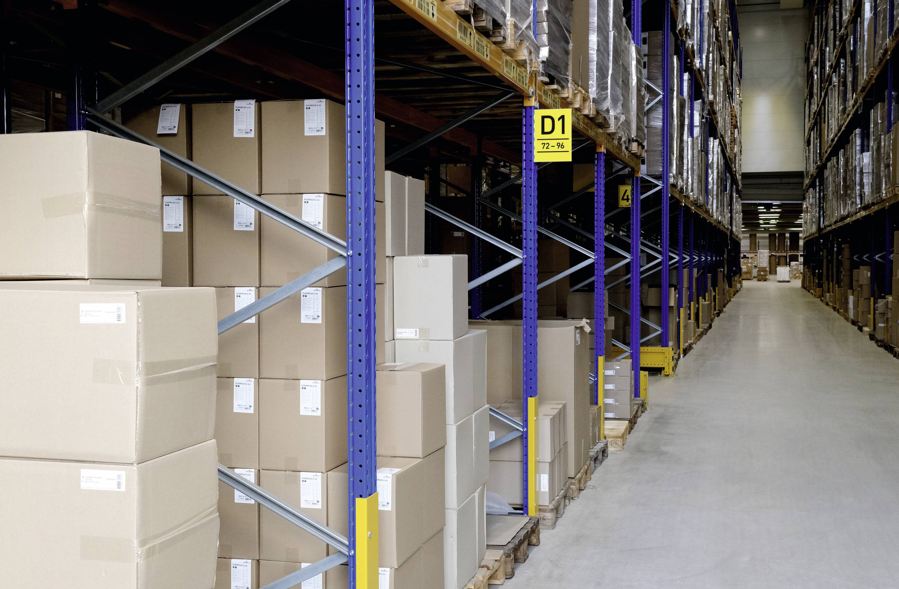 Large warehouse with stacked boxes on shelves. A long aisle separates the shelving units. A sign marks the shelf section as 'D1'.