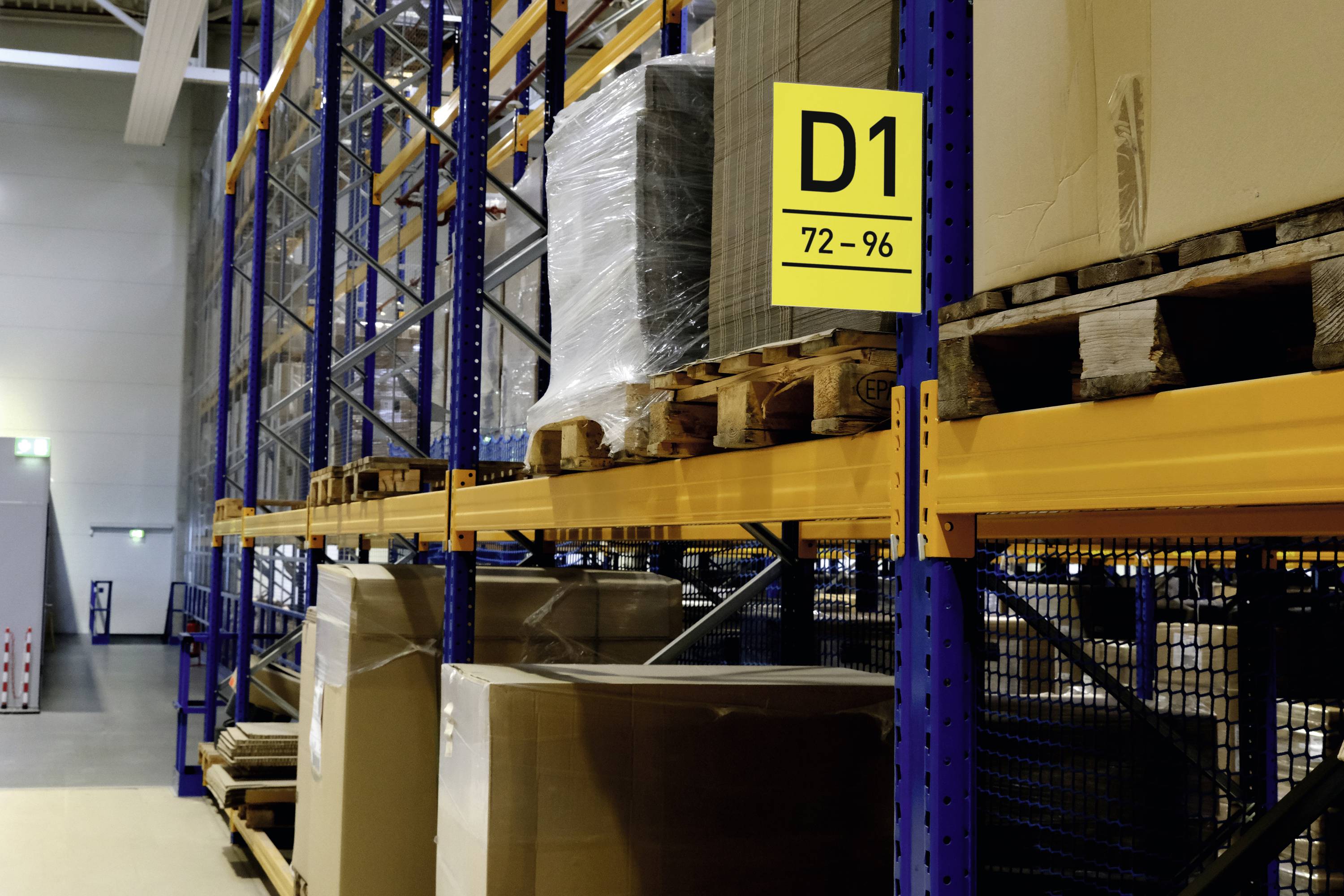 A storage rack with a yellow sign 'D1 72-96', with pallets and boxes stacked on it. Further shelving is visible in the background.