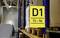 Yellow sign with the inscription 'D1 72-96' is hanging on a shelf in a warehouse. Pallets are stacked underneath.