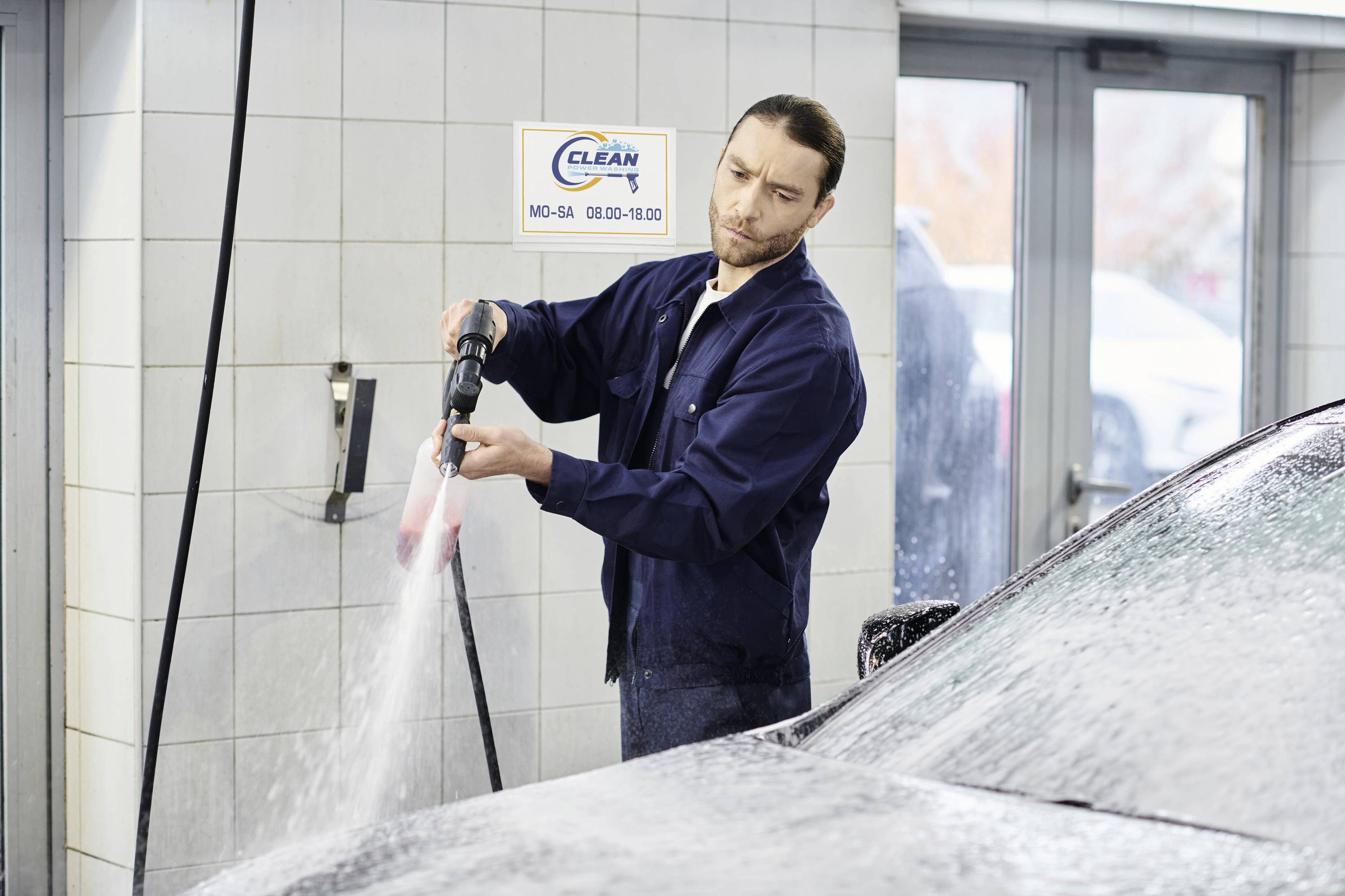 A man is cleaning a car with a pressure washer at a car wash. A sign with opening hours is hanging on the wall.