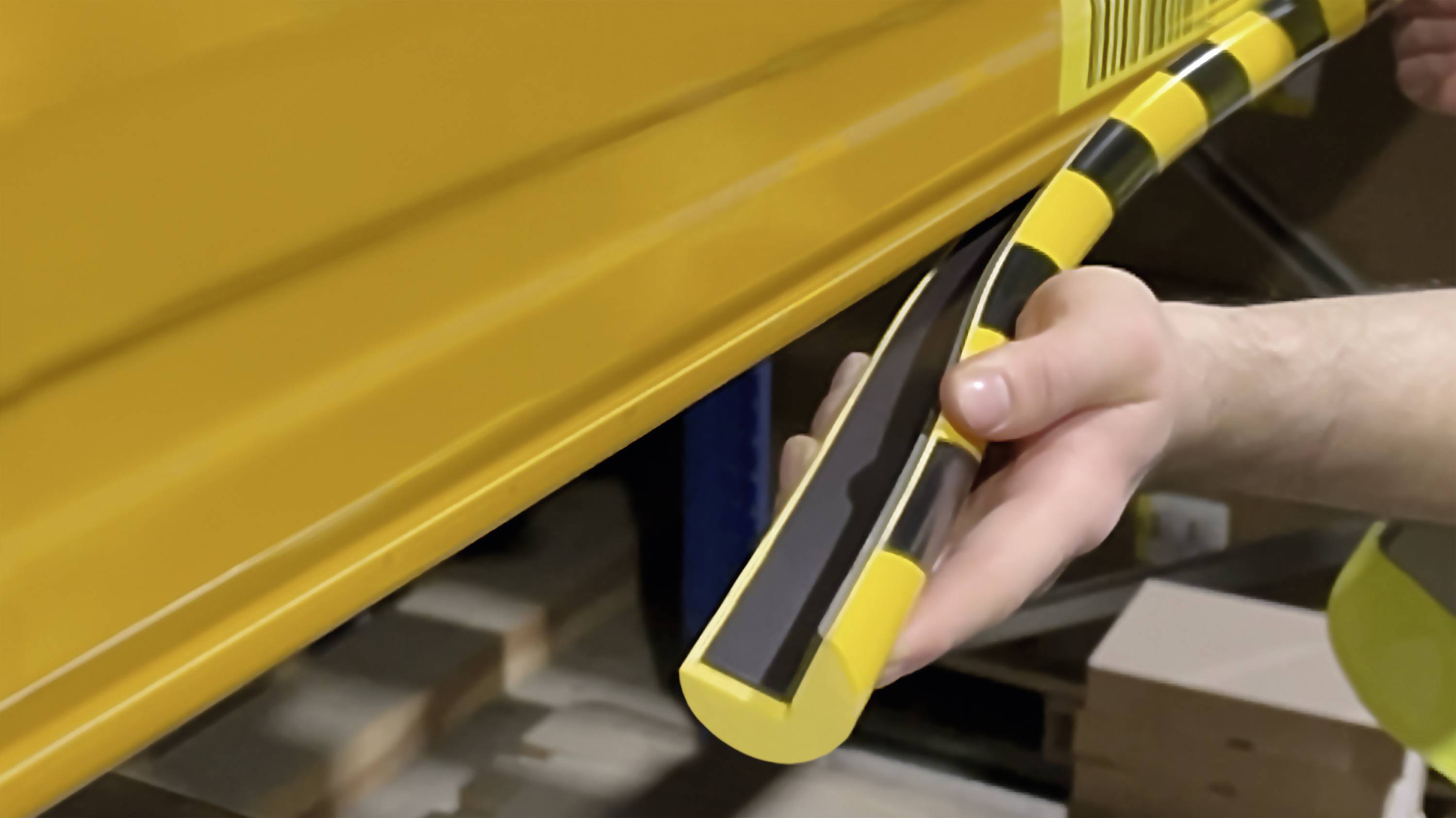 A person is applying a yellow protective profile with black stripes to a metal surface, likely for shock absorption or safety marking.