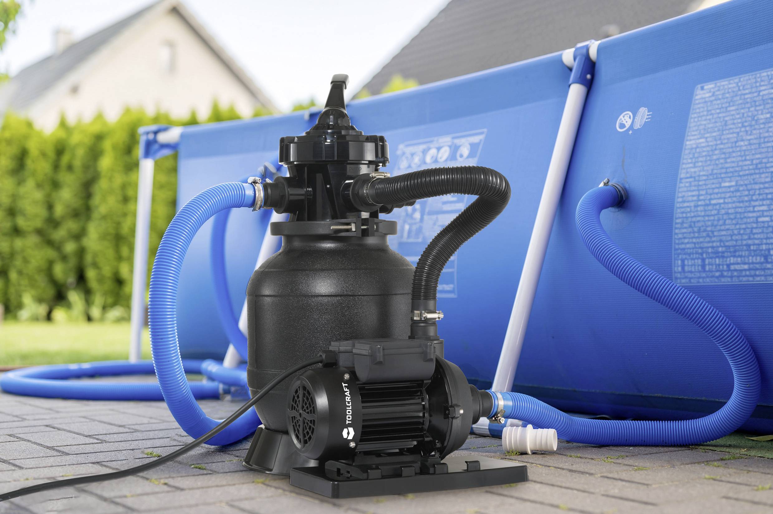 A sand filtration system next to a blue above-ground swimming pool, connected with hoses. It is situated on a paved outdoor surface.