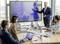 A businessman presents a chart with rising trends on a screen while four people listen attentively in a conference room.
