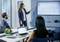 Woman presents data on screen in office. Two people listen, laptops open. Line graph shows rising trend.