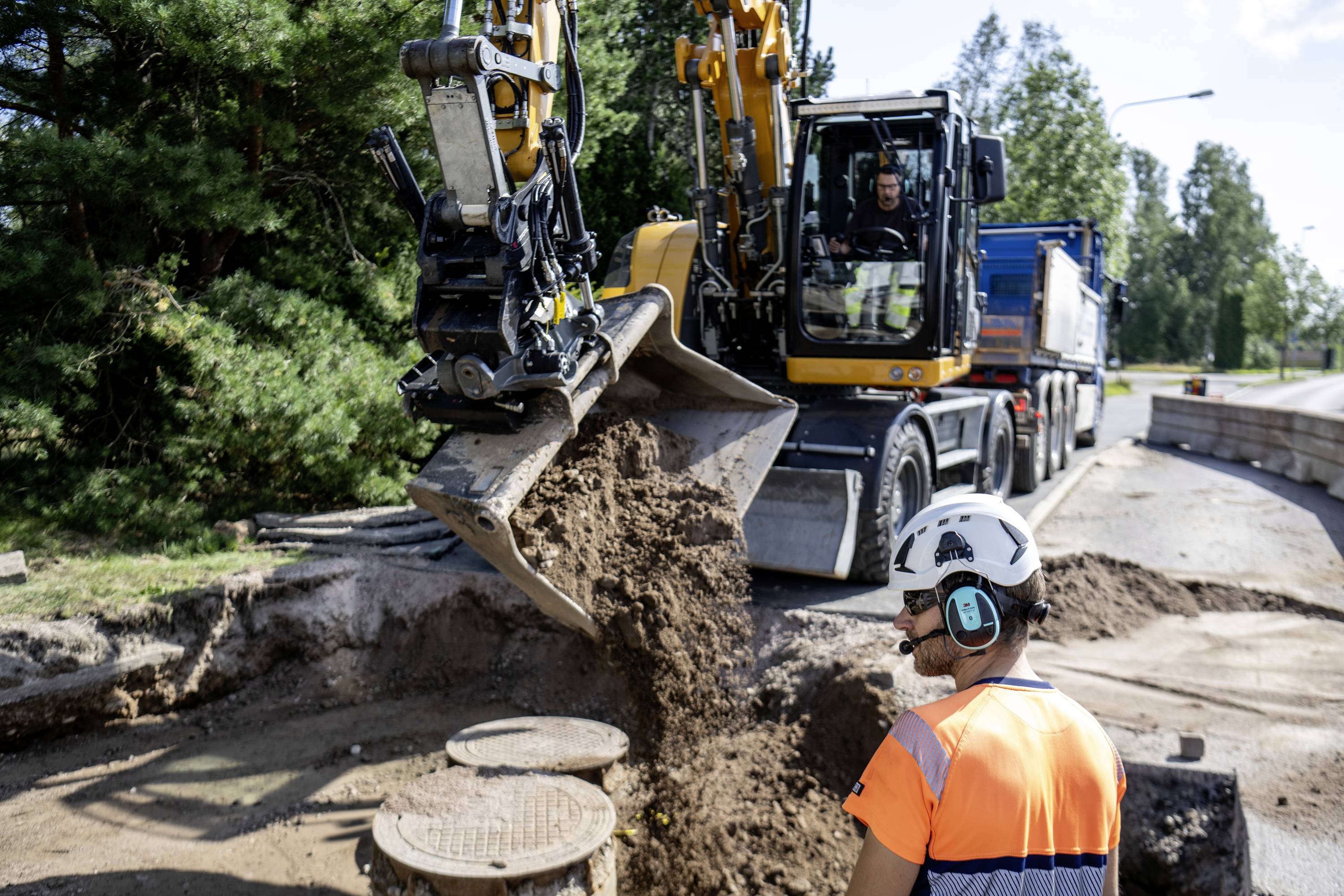 A construction worker wearing a hard hat and high-visibility vest is supervising a building site, while an excavator moves earth. A lorry is parked in the background.